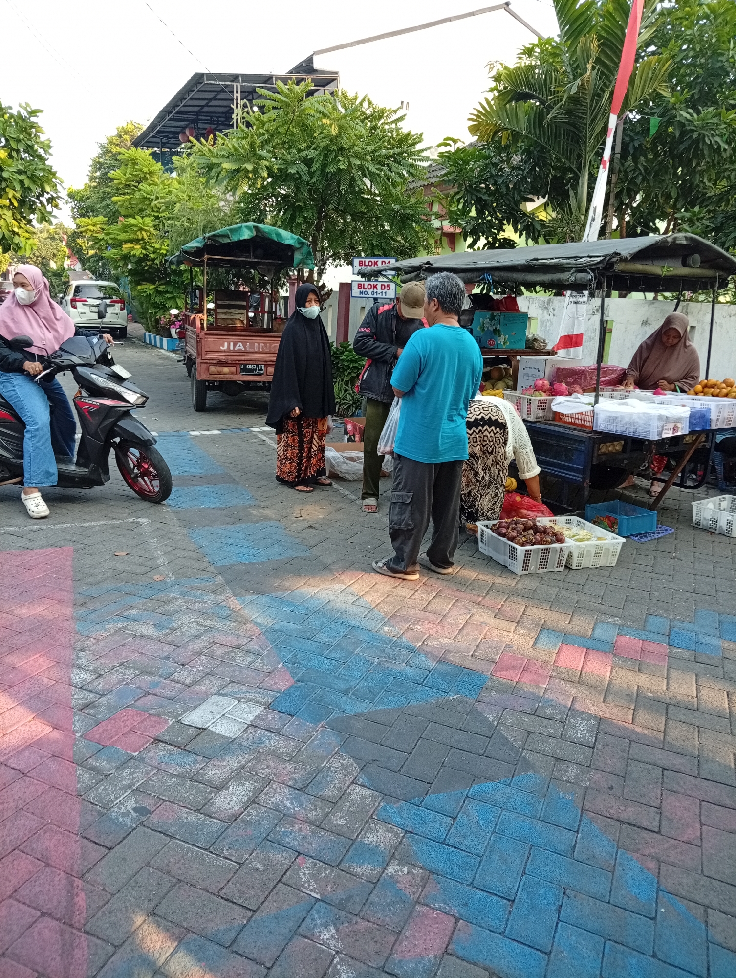 The image depicts a street scene in Bligo, Indonesia, with several individuals engaged in commerce. A woman wearing a pink hijab and face mask sits on a red and black Honda motorcycle. To her right, a woman in a black hijab and patterned skirt stands near a mobile food cart. Further to the right, another woman, also in a hijab, is seated behind a stall laden with fruit, including oranges and what appear to be dragon fruits and mangosteens. Two men are also present; one, wearing a blue t-shirt, faces away from the camera, while the other, in a beige cap and dark jacket, is partially visible. The ground is paved with interlocking bricks, marked with red and blue spray paint. Trees and buildings line the background, and a national flag of Indonesia is visible on a flagpole. The atmosphere suggests a local marketplace or roadside vending area.