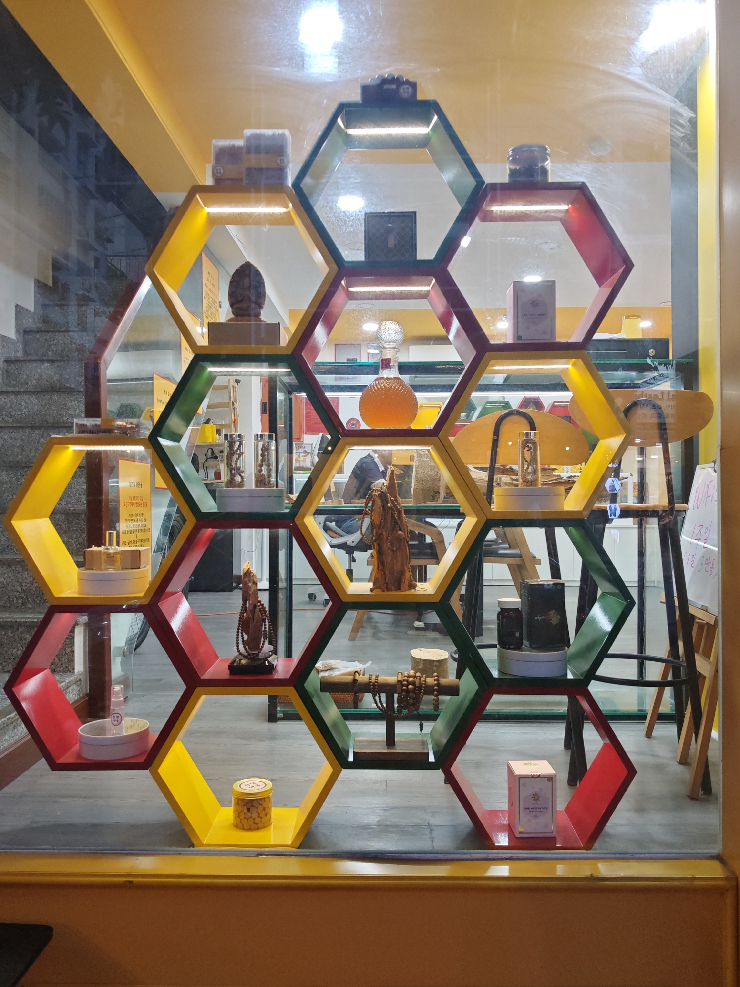 The image displays a shop window with a honeycomb-shaped shelving unit. The shelves are illuminated and contain various items including jars of what appear to be pills or seeds, boxes, a statue, and decorative objects. The shop appears to be located in Nha Trang Ward, Vietnam, indicated by the presence of Vietnamese text on a sign within the display. The overall scene is a retail display designed to showcase merchandise, possibly related to health or traditional remedies, given the items visible. The background shows a staircase and a reflective surface, suggesting the display is inside or behind glass.