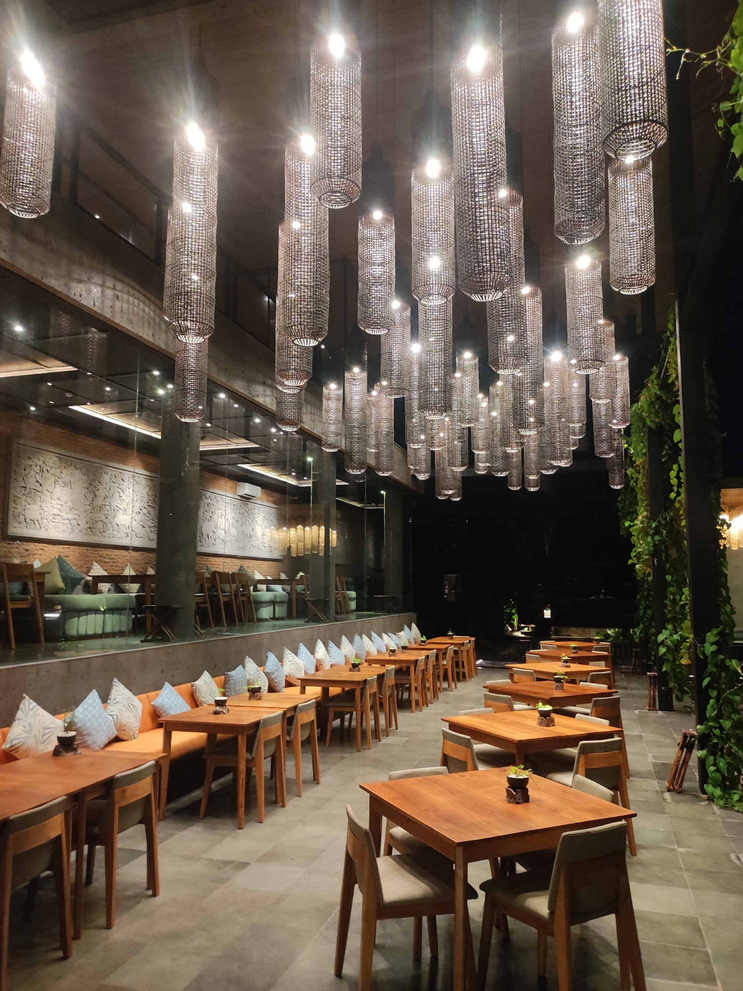The image depicts an unoccupied semi-outdoor dining area at night, located in Mas, Indonesia. The scene is illuminated by numerous cylindrical hanging lanterns with woven or slatted exteriors, casting bright light downwards from a high, exposed wooden ceiling.
To the left, separated by a reflective glass partition, is an indoor section featuring a long brick wall adorned with an extensive, white bas-relief sculpture depicting figures and scenes. Below the sculpture, an orange-cushioned bench seating runs along the wall, paired with individual wooden chairs and rectangular wooden tables. Light blue and white patterned pillows are arranged on the bench.
In the foreground and extending to the right, multiple rows of unoccupied rectangular wooden tables with matching wooden chairs featuring light gray upholstered seats are systematically arranged on a checkered tile floor. Each table features a small dark potted plant as a centerpiece. Beyond the dining area, the background is dark, indicating the nighttime environment, with lush green foliage climbing a structural column on the far right. No individuals are present.