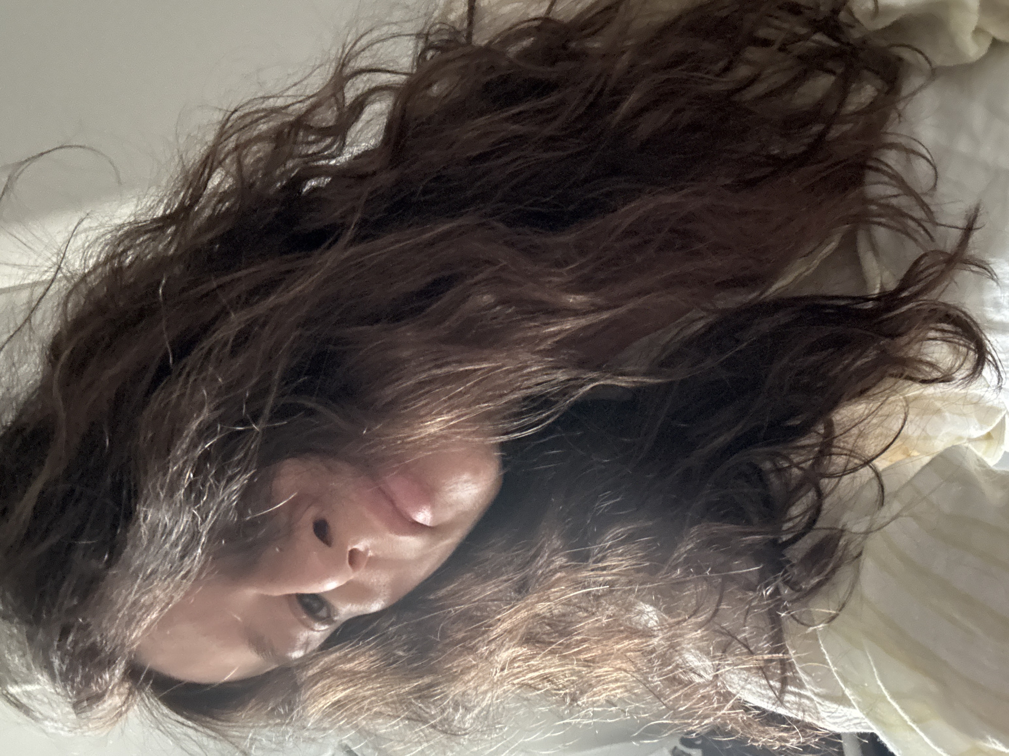 This close-up image, viewed from an overhead perspective, depicts a person with abundant, dark brown, wavy hair spread around their head. The hair appears somewhat disheveled, suggesting a relaxed state. The person's face is partially visible, showing the bridge and tip of the nose, the area around the mouth, and one eye. The visible eye is open, and the expression appears neutral.
The individual is likely lying down, possibly on a bed or a soft surface, given the informal posture and spread of hair. To the right and below the person's head, a light-colored, possibly cream or off-white fabric with subtle vertical stripes is visible. This fabric could be a bedsheet, pillowcase, or an item of clothing, indicating an indoor setting, most likely a bedroom.
The lighting is soft and diffused, without harsh shadows, consistent with ambient light from a window during the daytime or early morning/late evening. No specific time of day or weather conditions can be definitively determined. While the general setting suggests an indoor space, there are no specific location cues within the image itself to confirm the provided context of Chaikovsky, Russia. The overall mood is intimate and calm. There is no visible text in the image.