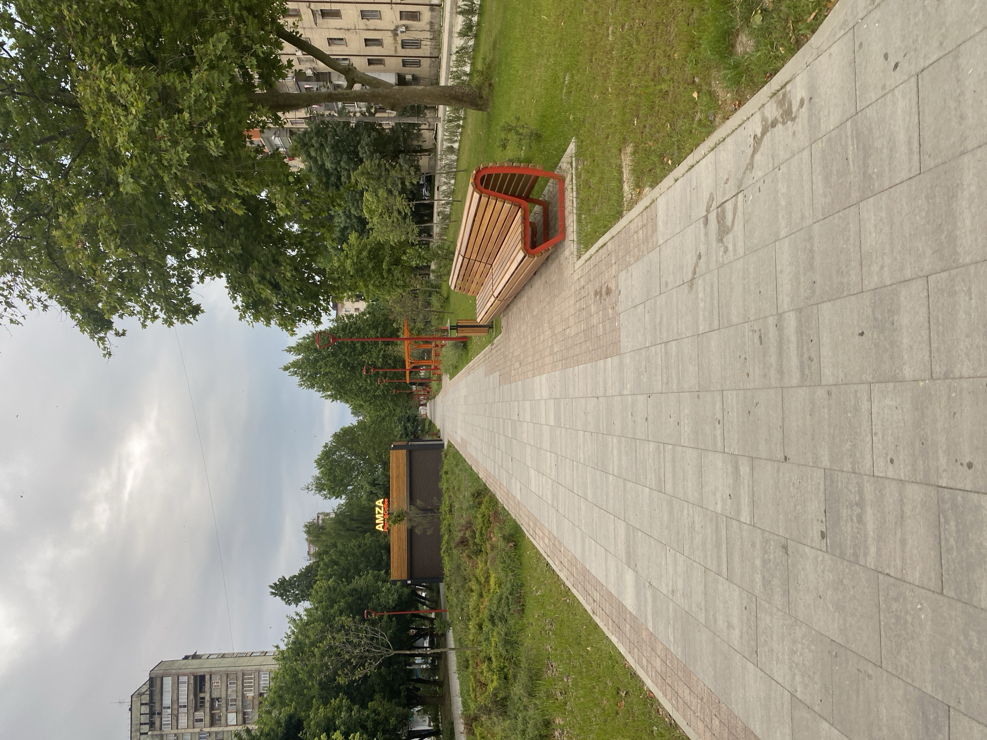 The image depicts a paved walkway in an urban park. The walkway is lined with green grass and trees on both sides, with buildings visible in the background. A modern wooden bench with red accents is positioned on the right side of the walkway, facing away from the camera. Further down the walkway, a small structure with an illuminated "AMZA Pizza & Coffee" sign is visible. The sky is overcast, suggesting a cloudy day. There are no people or animals visible in the scene. The overall impression is one of a well-maintained public space.