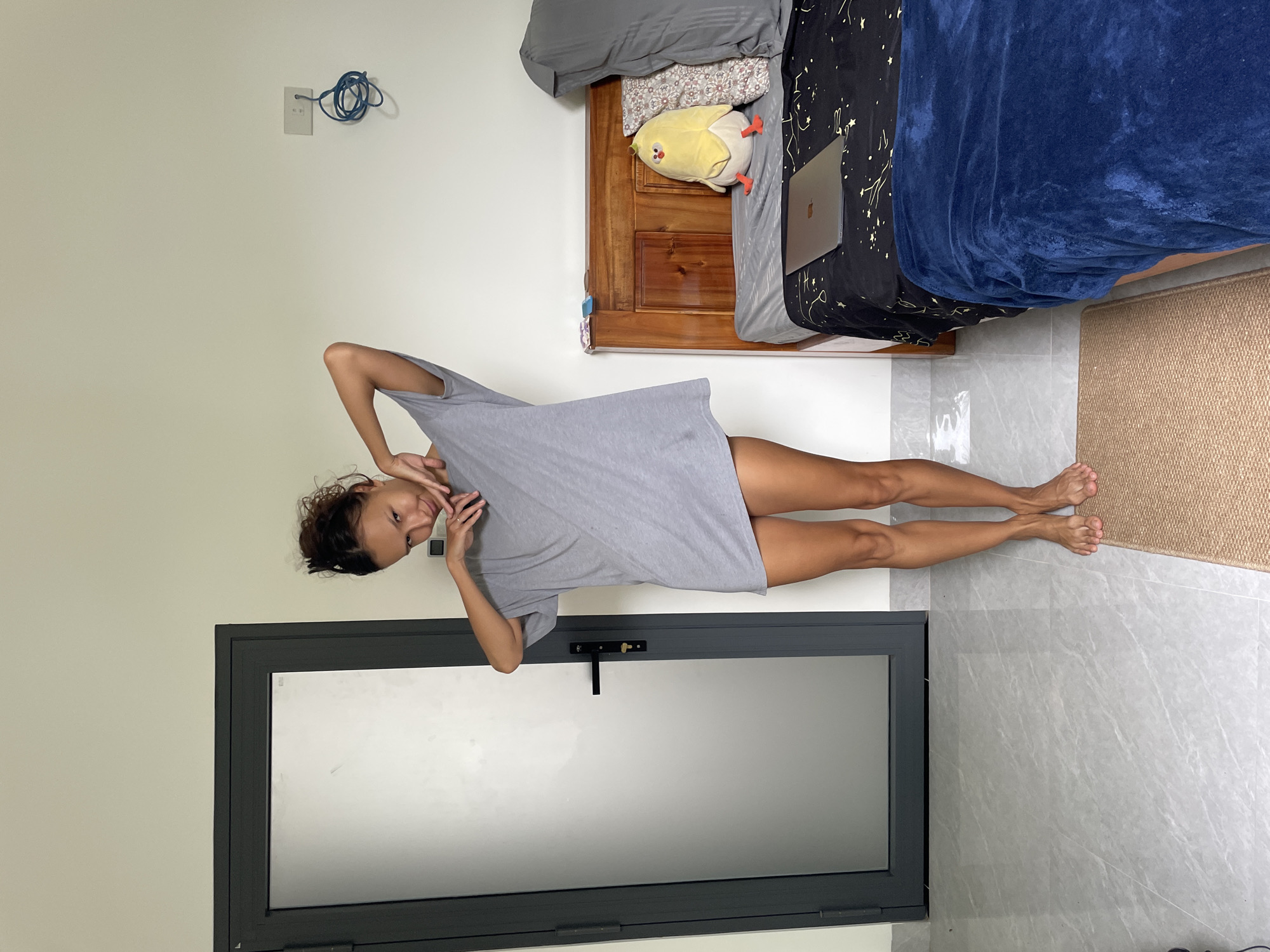 A young woman is captured posing playfully in a brightly lit indoor room, likely a bedroom or hotel room, in Mũi Né, Vietnam. She stands with her body slightly turned, her left hand near her face, and her right arm raised towards her head, looking towards the camera with a relaxed smile. She has dark, curly hair tied up and is wearing a loose-fitting, light grey t-shirt or dress that reaches mid-thigh, revealing her tanned legs and bare feet.
To the right of the woman, a wooden bed frame is made with grey bedding, a patterned pillow, a black duvet cover featuring golden star and constellation designs, and a blue fuzzy blanket draped across it. A silver laptop, possibly a MacBook, is open on the bed, and a yellow chick-shaped plush toy sits near the headboard. On the upper left wall, an electrical outlet with blue coiled wires is visible. A large, grey-framed glass door or window, equipped with a black handle and latch, occupies the lower left portion of the image. The room features plain white walls and a floor covered in large grey marbled tiles. A small, light-colored woven mat or rug is placed on the floor beside the bed. The scene suggests a casual and cheerful moment. No visible text is clearly discernible.