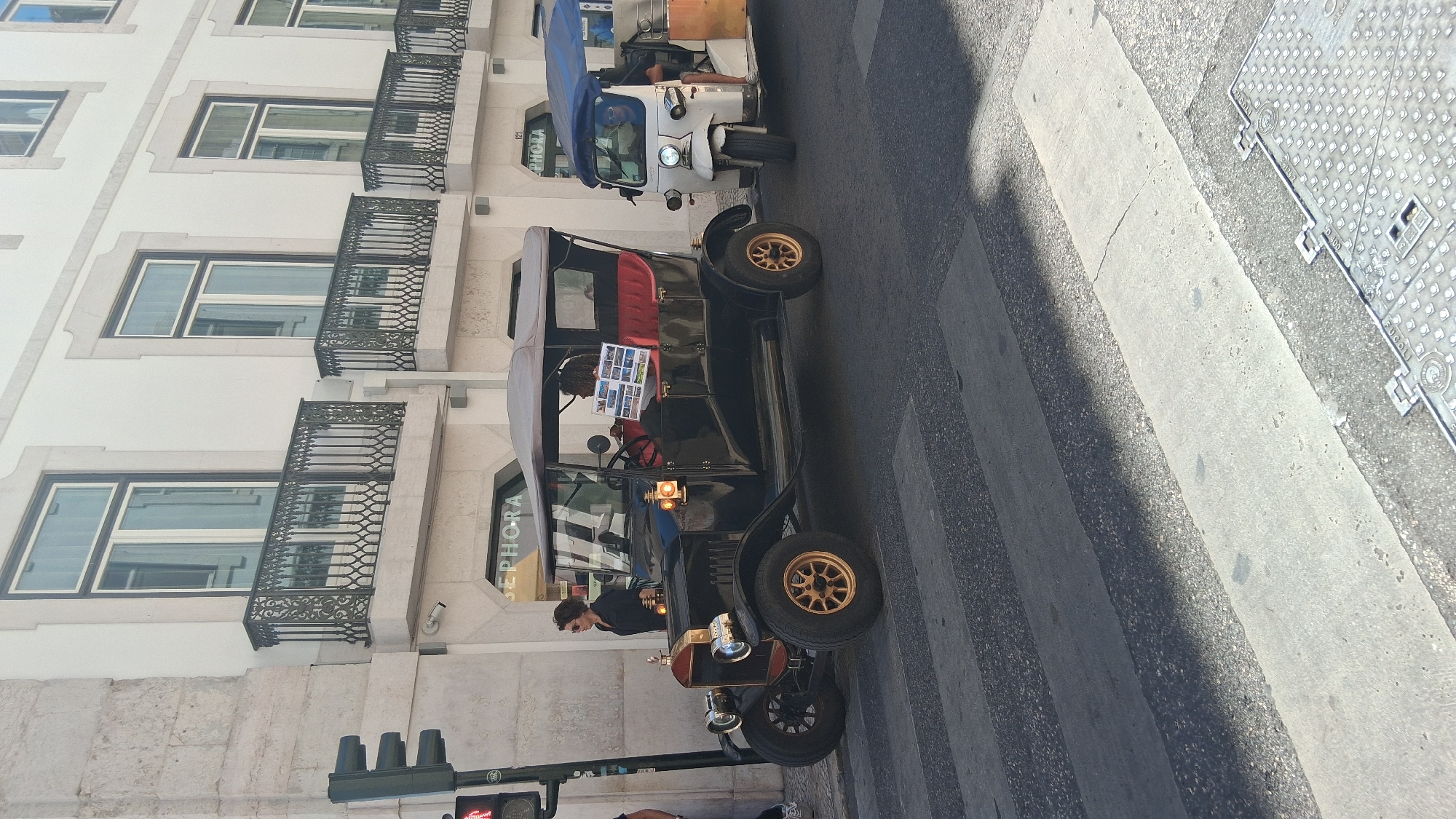 A vintage black car with red interior and a tan convertible top is stopped at a crosswalk in front of a building with "SEPHORA" signage. A woman in a black shirt and sunglasses stands on the sidewalk to the left of the car. To the right of the vintage car, a white three-wheeled tuk-tuk with a blue canopy is also stopped. The tuk-tuk has a driver wearing sunglasses. The scene takes place on a street in Lisbon, Portugal. The building has ornate black metal balconies above the storefronts.