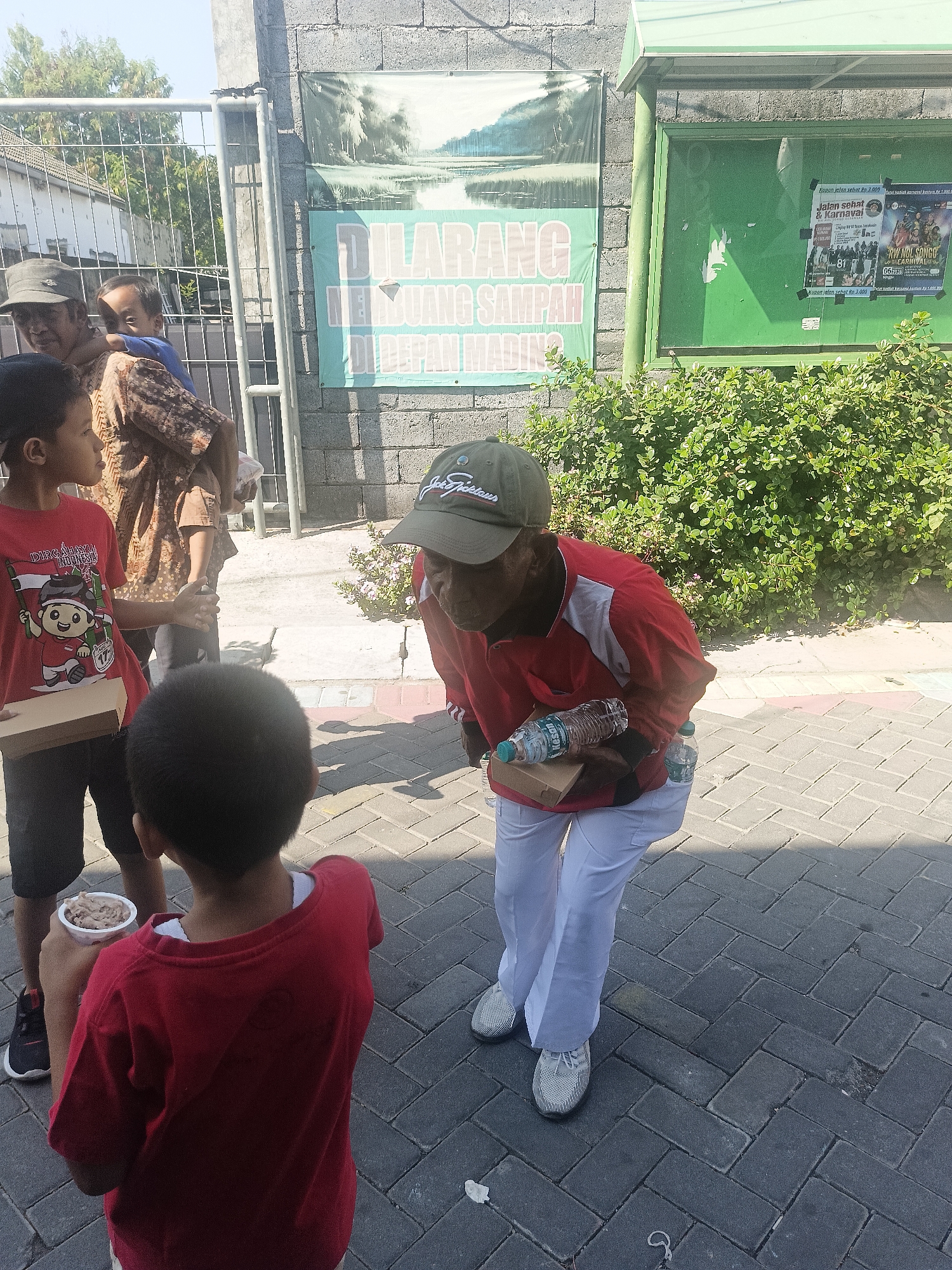 In Gedangan, Indonesia, on a sunny day, a man wearing a red and white shirt, a green cap, and white pants is standing on a paved walkway. He is bent over and appears to be offering or handing something to a child. He is holding two plastic water bottles and a small brown box.
Several children are present. One child, wearing a red t-shirt and black shorts, stands facing away from the camera, holding a cup with what appears to be food in it. Another child, wearing a red t-shirt, is visible from the side, and a third child, being held by an adult, is also in the frame. An adult, wearing a patterned brown and white top and a cap, is holding the child.
Behind the group, a brick wall is visible with two large signs. One sign has a picture of a natural landscape and the words "DILARANG MEMBUANG SAMPAH DI DEPAN MASJID" (Do not litter in front of the mosque). Another sign, on a green board, displays several posters. The surrounding area includes a metal fence, some green bushes, and more paved walkway. The lighting suggests it is daytime with a clear sky.