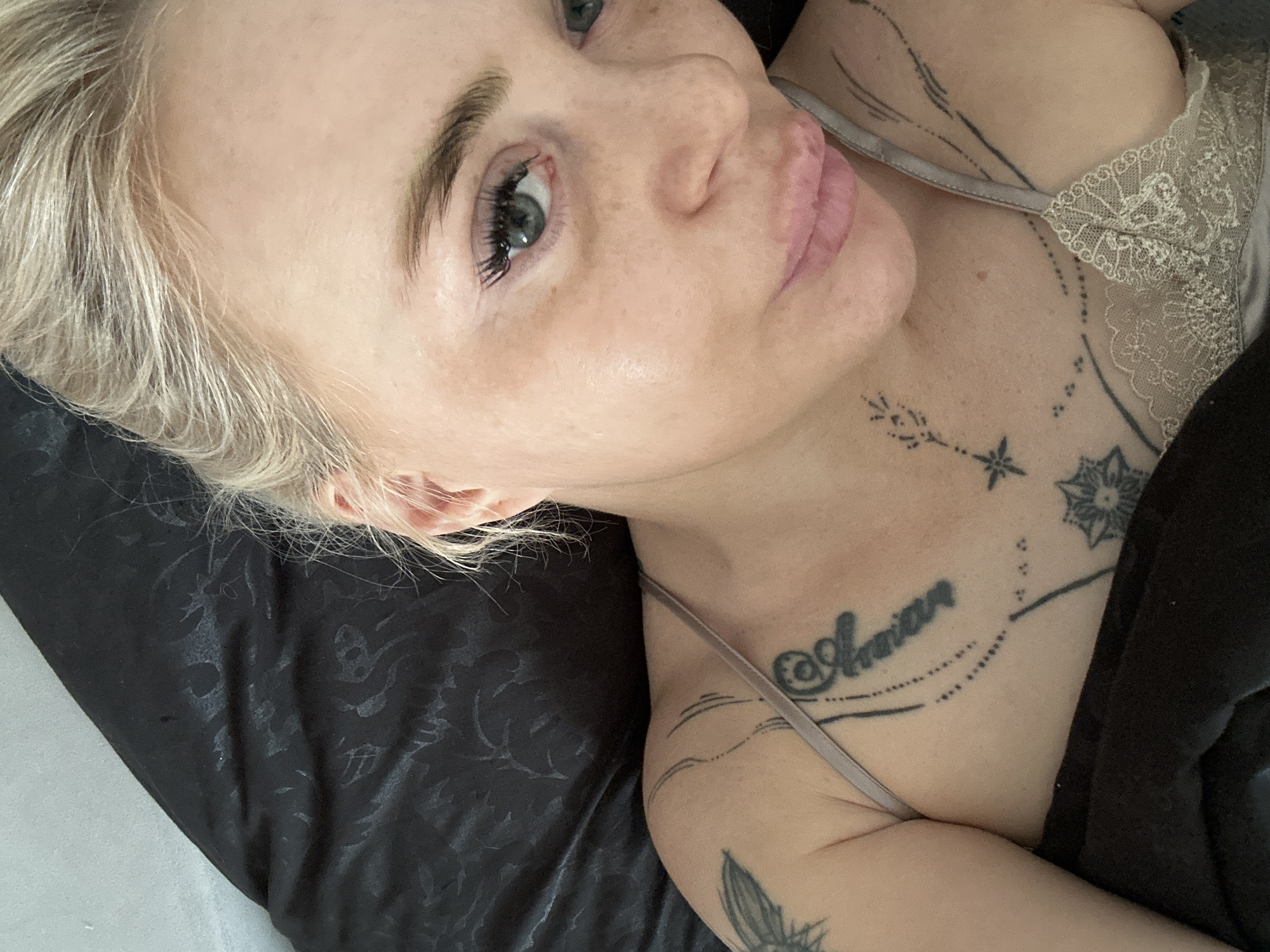 A close-up, high-angle selfie depicts an adult female with blonde hair and fair skin, lying on dark bedding. Her head rests on a black or very dark grey pillow with a subtle, interwoven pattern, possibly satin or silk. Her blonde hair is parted to the side, with some strands visible near her temples and ears. She looks directly at the camera with blue-green eyes, which feature prominent dark eyelashes. Her eyebrows are light brown and defined. Her lips are full, light pink, and slightly parted. Her skin shows some visible sheen, particularly on her forehead and cheekbones, and some subtle hyperpigmentation or discoloration below her eyes.
She is wearing a light, possibly beige or champagne-colored, camisole or bralette with thin straps and lace detailing on the cups. Multiple black ink tattoos are visible on her chest and shoulders: an intricate design featuring script (partially visible, possibly "@Amira"), dotted lines, and geometric shapes covers her upper left chest; a delicate floral or starburst pattern is on her lower left chest; a small, star-like design with branching elements is on her left clavicle; and a larger, abstract, wing-like or feather design is on her upper right shoulder near the armpit.
The background consists primarily of the dark patterned bedding, with a small portion of a lighter, possibly off-white, wall or headboard visible in the upper left corner. The lighting appears soft and even. The location is in the city of Unknown, Unknown.