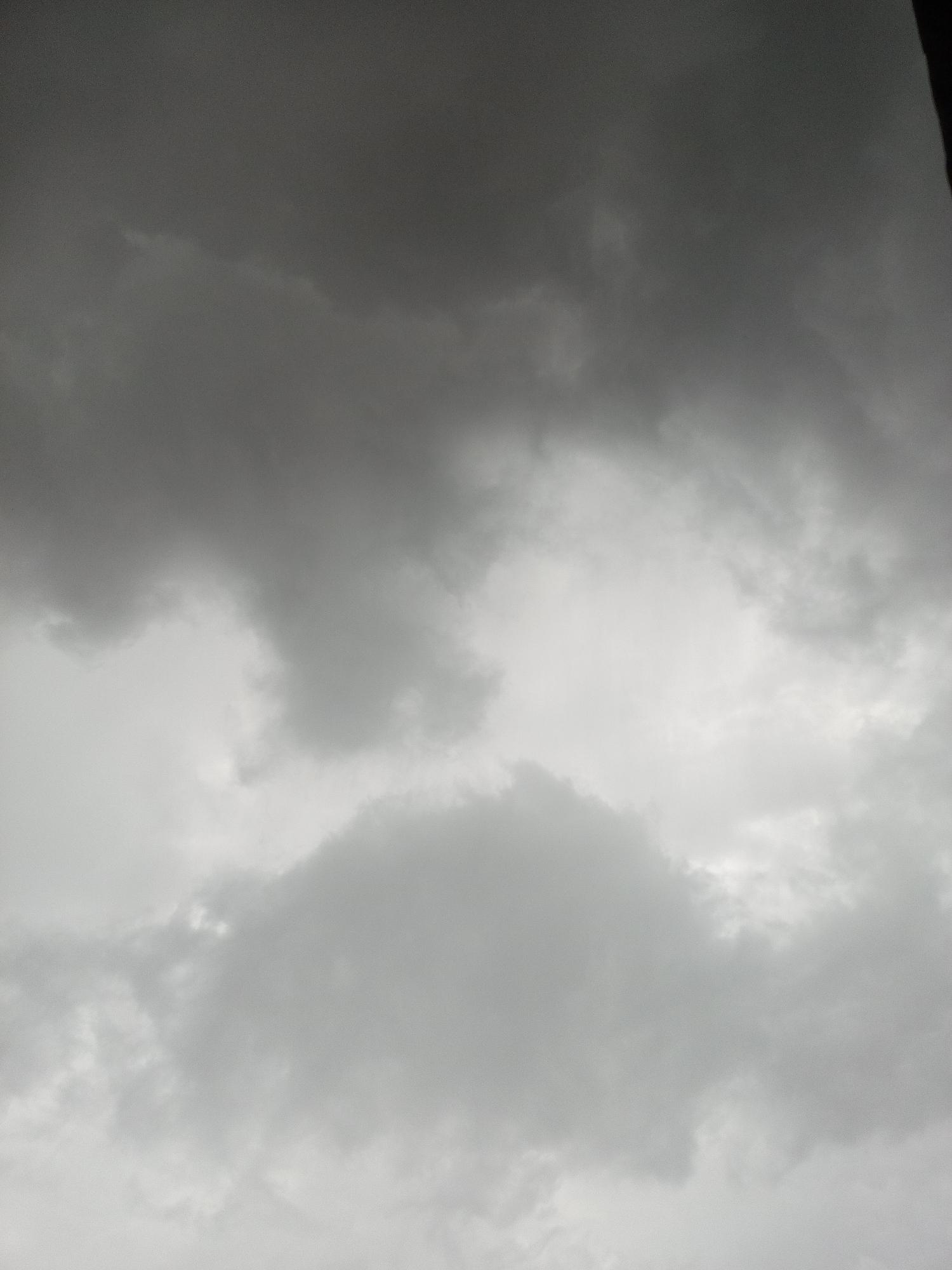 The image shows a sky filled with dark, ominous storm clouds. The clouds are a mix of grays and blacks, suggesting an impending downpour or an ongoing storm. There is a hint of lighter gray in the center of the image, possibly where some light is breaking through, but the overall impression is one of overcast and turbulent weather. The perspective is looking upwards, with no visible ground or man-made structures.