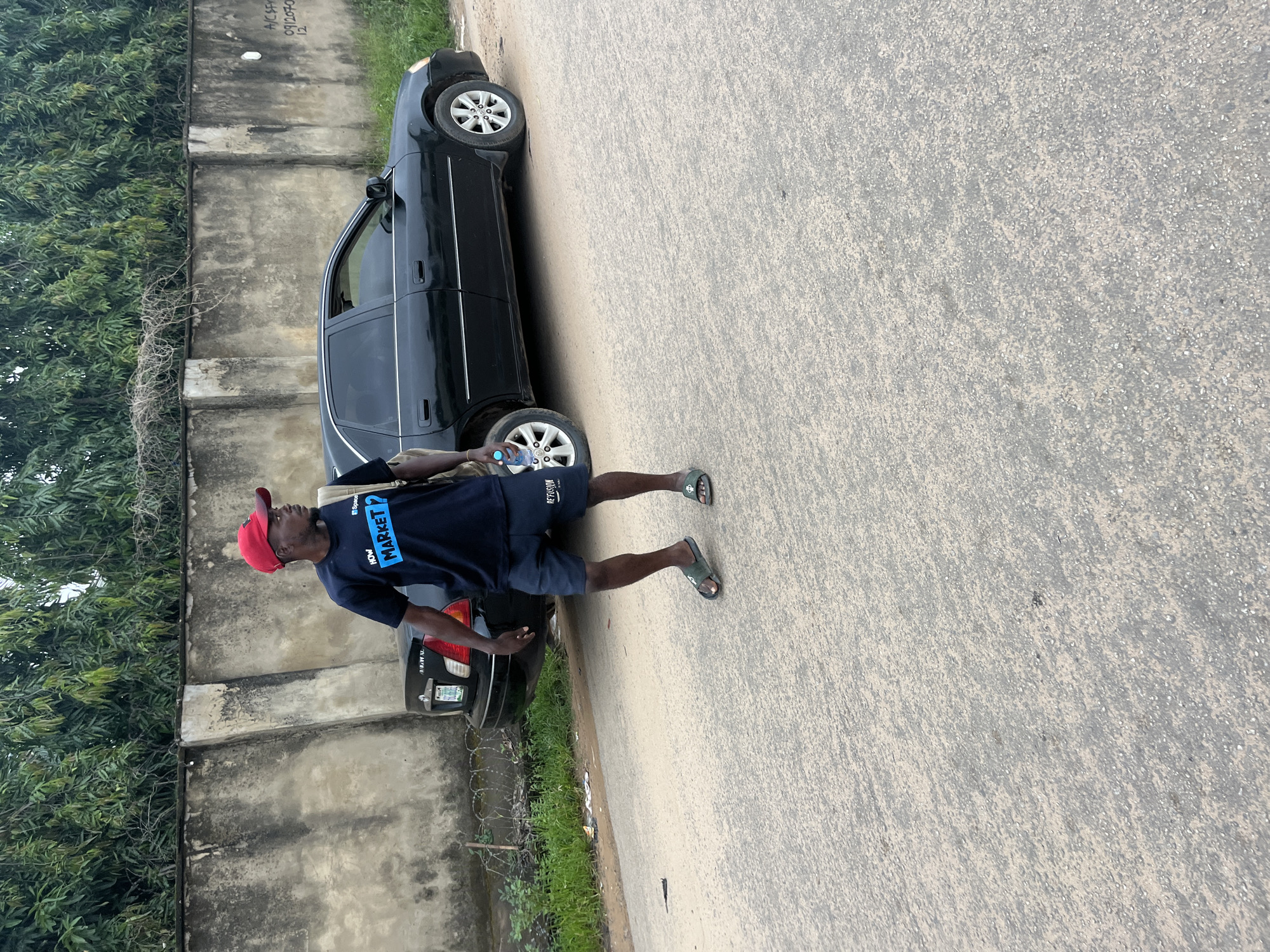 A dark-skinned man wearing a red baseball cap, a dark blue t-shirt with blue text reading "HOW MARKET?", and dark blue shorts stands on a paved road. He is carrying a light brown bag over his left shoulder and holding a clear plastic bottle of water in his right hand. He is looking to his left. Behind him is a black sedan. A textured concrete wall with some graffiti runs along the background, topped with barbed wire and a dense green foliage of trees. The scene is likely located in Abuja, Nigeria, given the context.