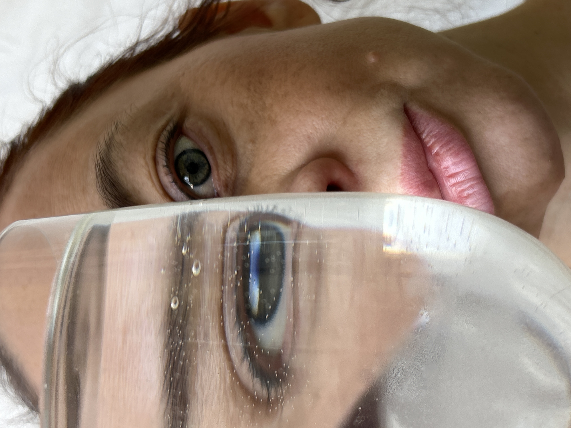The image shows a person's face, partially obscured by a clear, likely cylindrical, object. The upper portion of the face, including an eye and part of the nose and mouth, is visible above the object. A distorted, refracted reflection of the lower part of the face, including an eye, is visible through the object, which appears to have some condensation or droplets on its surface. The background appears to be a white or light-colored surface. No information about Chaikovsky, Russia is discernible in the image.