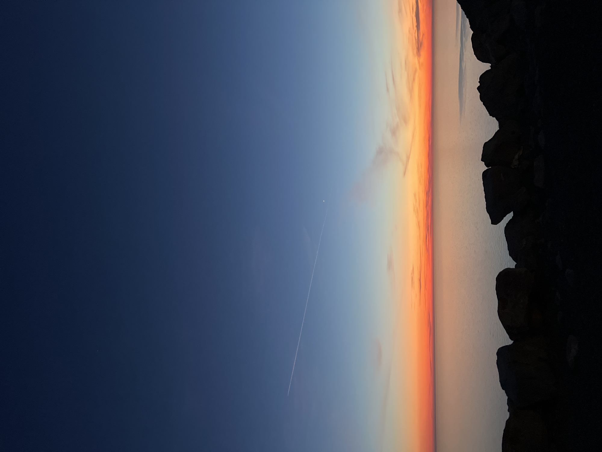 The image captures a sunset at Kovačko Polje, Montenegro. Dark, silhouetted rocks are on the right side of the image, bordering the water. The water reflects the fading light of the sunset. The sky fades from deep blue on the left to orange and pink hues near the horizon. A contrail from an aircraft is visible streaking diagonally across the sky. A bright star is also visible in the dark blue sky.