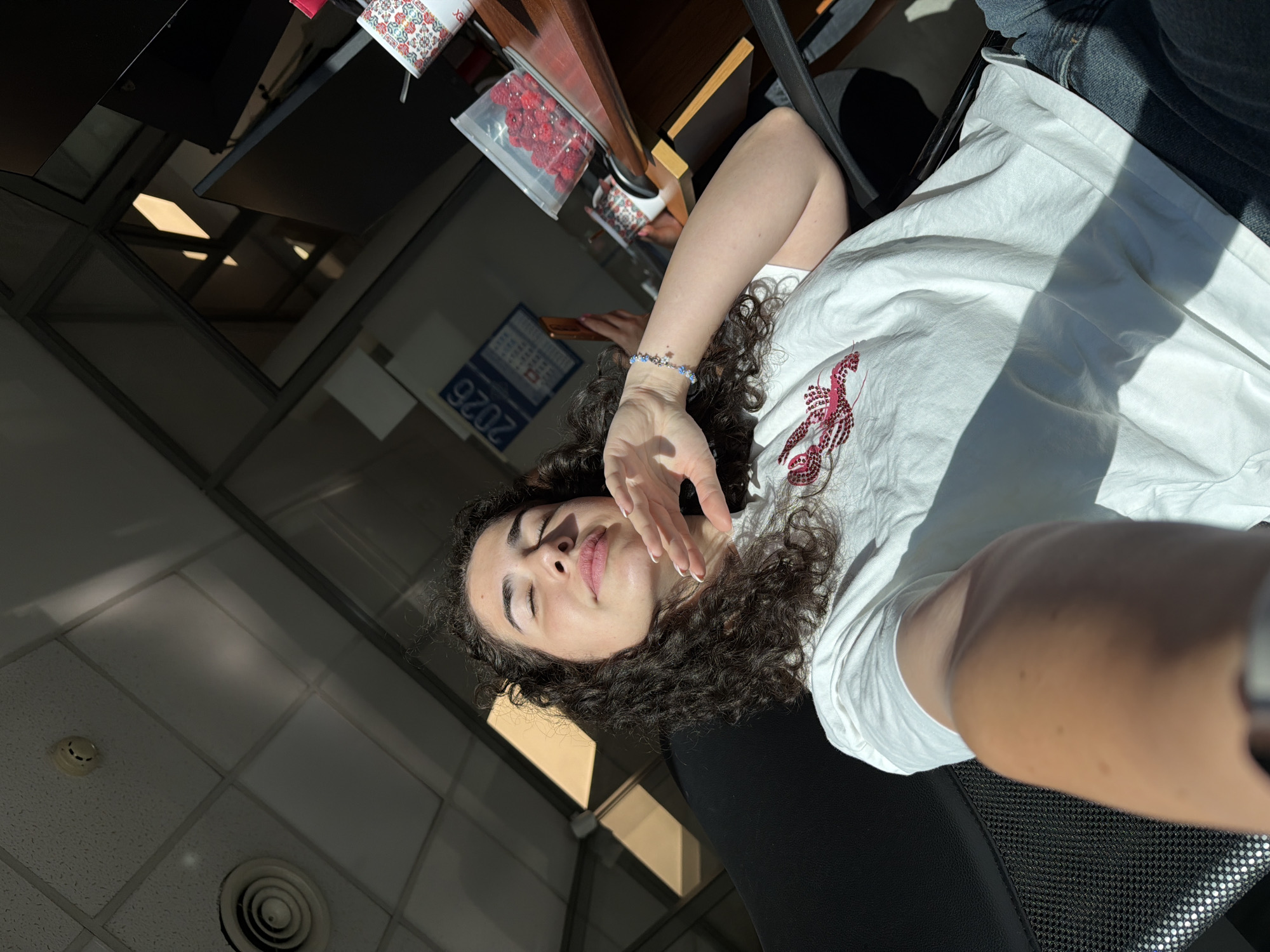 In the city of Sofia, Bulgaria, an outdoor selfie is captured, where a young woman with curly dark hair is positioned in the foreground. She is wearing a white t-shirt with a red lobster graphic and blue jeans. Her eyes are closed, and her right hand is raised to her face, with her left arm resting on her lap. The background reveals a ceiling with fluorescent lighting and air vents, suggesting an indoor environment. A calendar with the year "2026" is visible on the wall, along with a transparent container filled with red berries on a wooden desk. The scene is illuminated by natural light streaming in from the side, casting shadows across the room and the woman.