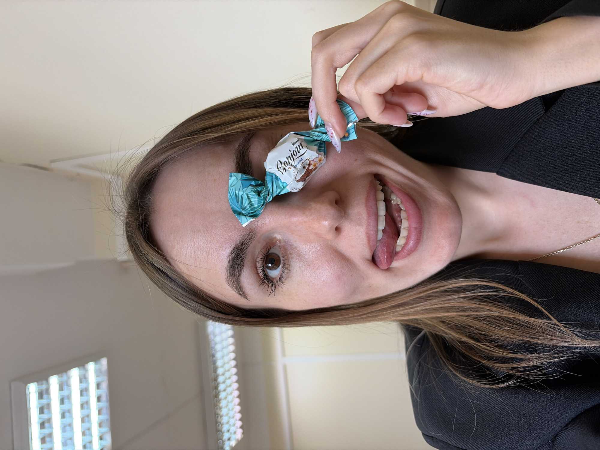 A woman holds a Bonjour coconut candy in front of her face, obscuring one eye. She is indoors, possibly in an office setting, as indicated by the fluorescent lighting panel in the background. The woman has brown hair and is wearing a black blazer. Her tongue is slightly extended. The location in the city of Izhevsk, Russia is not discernible from the visual information provided.