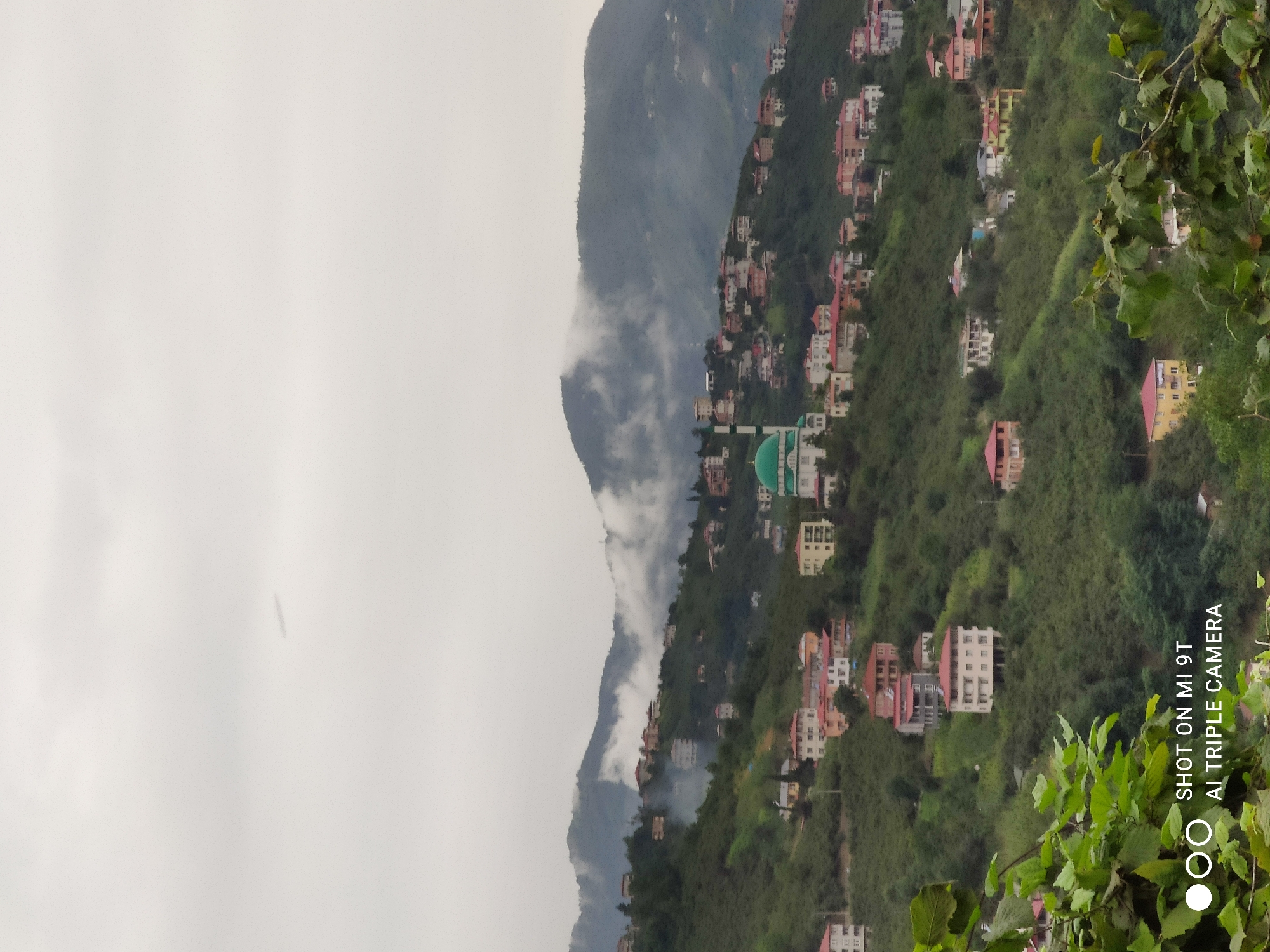 A panoramic, slightly overcast view captures a mountainside village near Akçaabat, Turkey. Numerous multi-story houses with red and white tiled roofs are scattered across the lush green, undulating terrain. A prominent mosque with a green dome and minaret is situated on the left side of the frame. The sky is a muted gray, with wisps of fog or clouds clinging to the distant mountain peaks. In the foreground, the leaves of lush green trees partially obscure the lower portion of the image. A watermark in the bottom left corner reads "SHOT ON MI 9T AI TRIPLE CAMERA." There are no people visible in the scene.