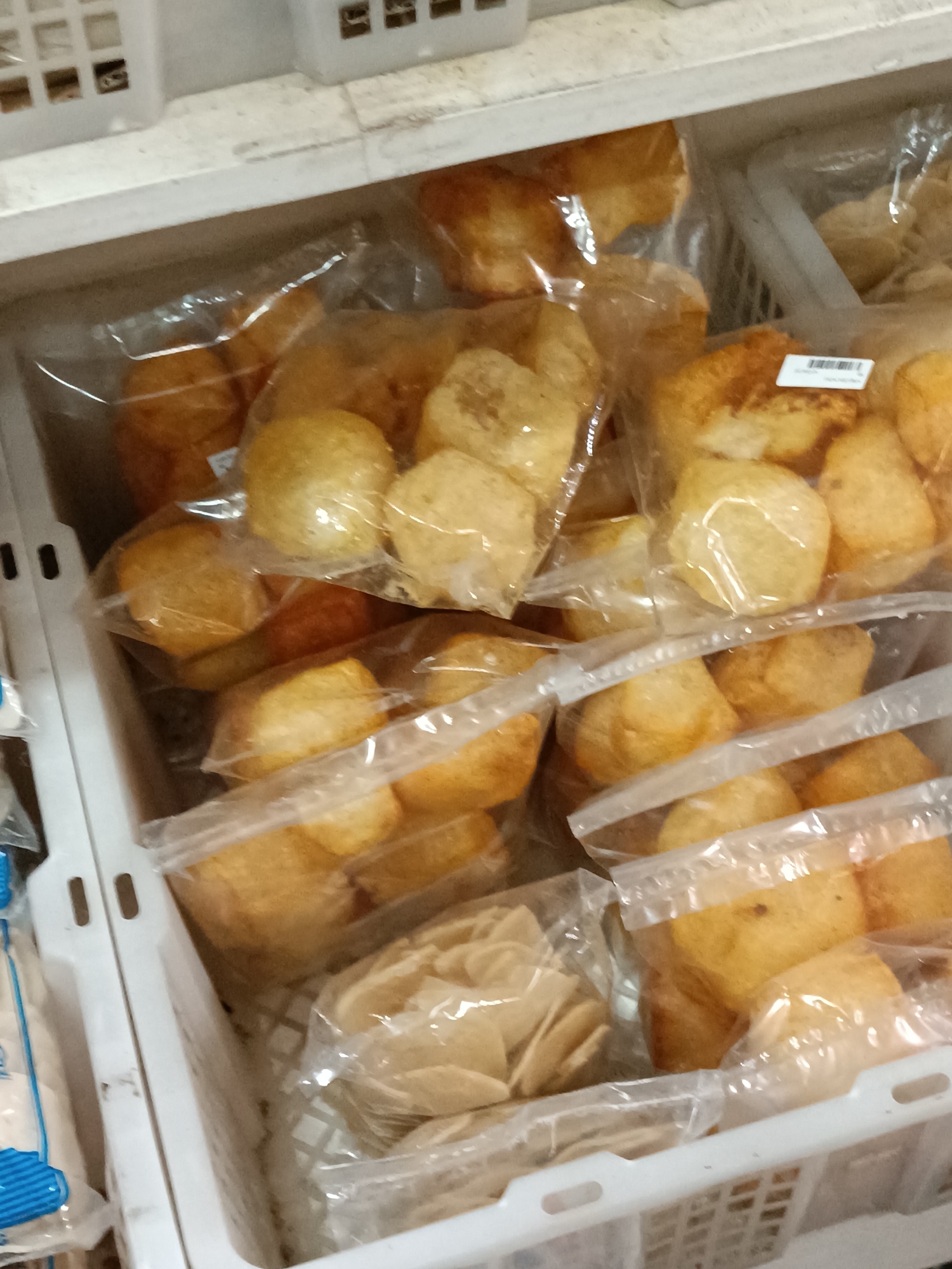 In the city of Jetis, Indonesia, within a store or market setting, a white plastic basket is filled with various food items packaged in clear plastic bags. The primary contents are circular, golden-brown fried dough balls, possibly a type of fritter or pastry, which are individually or loosely bagged. In the foreground, there is also a bag containing a stack of flat, light brown, possibly crispy food items, arranged in a fan-like shape. The basket is positioned on a shelf, with other similar baskets visible above it, suggesting a retail display. The lighting is ambient, and the image is taken from a slightly elevated angle, looking down into the basket.