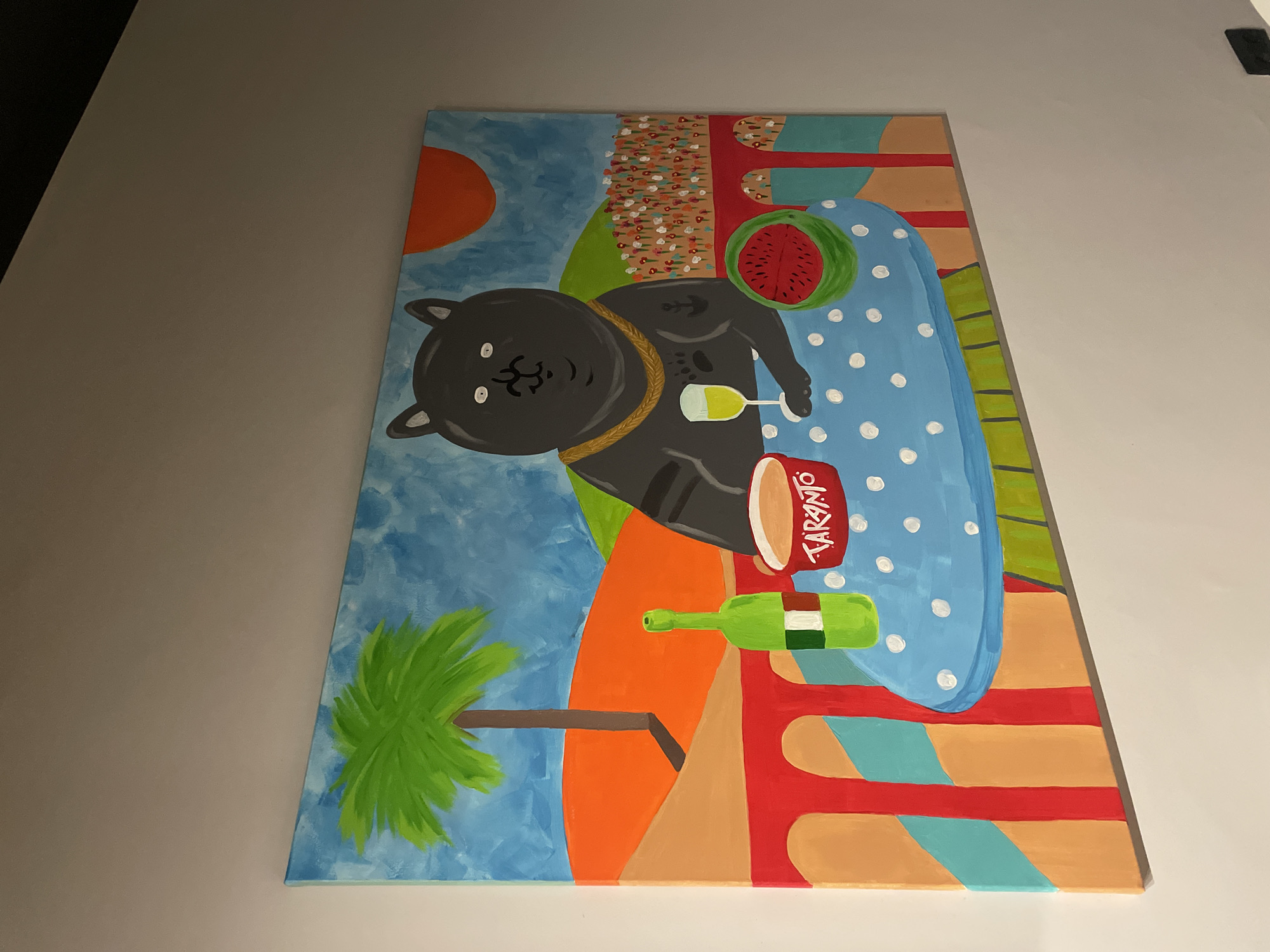The image shows a painting on a canvas. The painting depicts a black cat with a gold chain lying on a blue tablecloth with white polka dots. Next to the cat are a glass of wine, a watermelon, a bottle of wine, and a container labeled "TARANTO". The background includes a stylized landscape with a blue sky, an orange sun, a beach with people, a palm tree, and colorful geometric patterns. The location is not evident in the image.