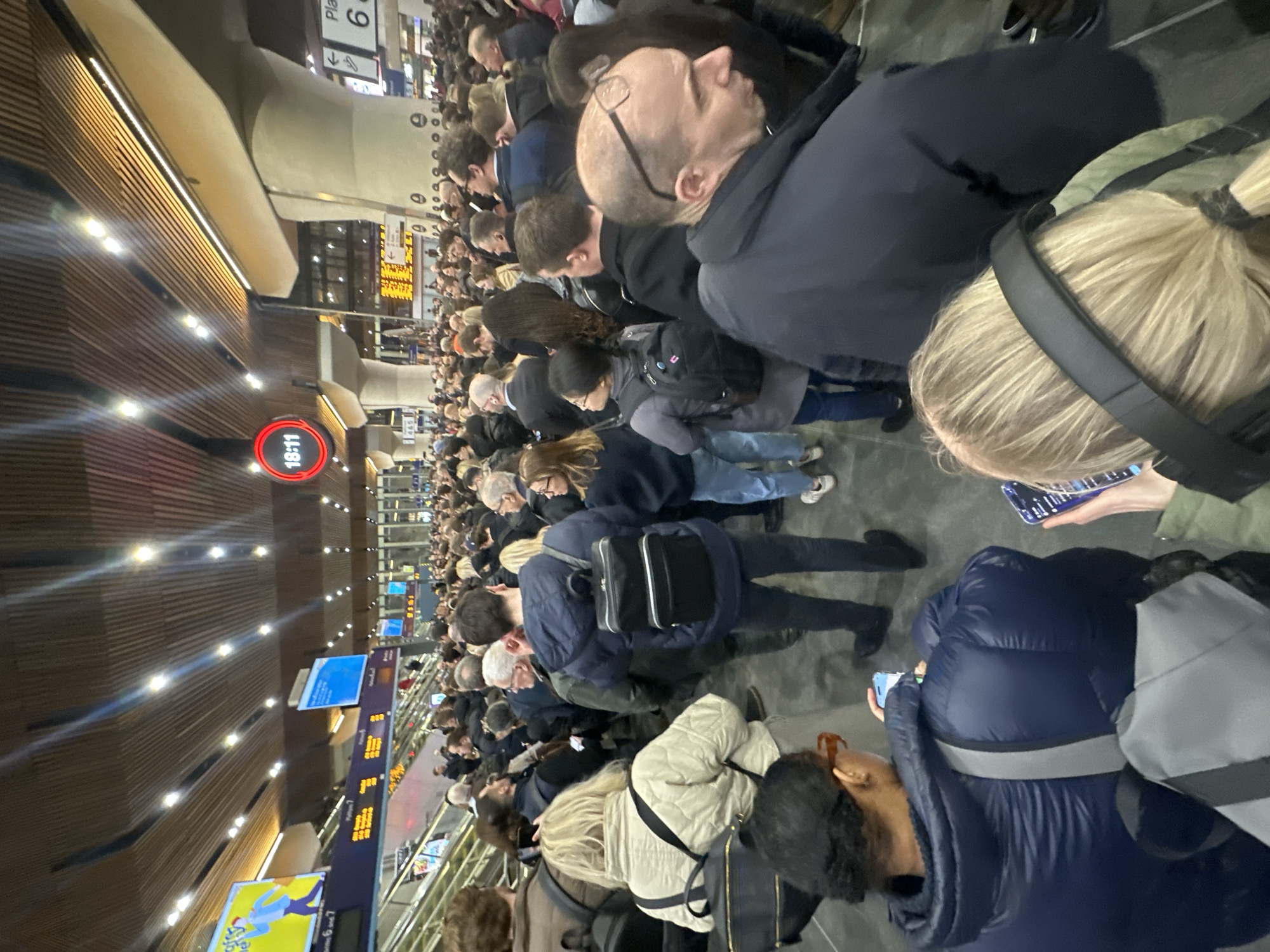 This image depicts a severely congested concourse at London Bridge Station, London, United Kingdom, at approximately 18:11, as indicated by a prominent circular digital clock. A large crowd of individuals, predominantly adults, fills the wide-angle frame, appearing densely packed. Most people are standing still, facing generally away from the camera and towards the center or right of the frame, suggesting a bottleneck or waiting area. Many individuals are observed holding and looking at smartphones. Numerous people are equipped with backpacks. The environment features a distinctive high, slatted wooden ceiling with integrated strip and spot lighting, supported by large, smooth, light-colored concrete pillars. In the background and to the left, escalators are visible, along with multiple digital display boards; one large board prominently displays "Platform 6" and other train information in yellow text on a dark background. Another screen displays an advertisement. The overall scene suggests a busy rush hour or a service disruption causing a significant accumulation of passengers.