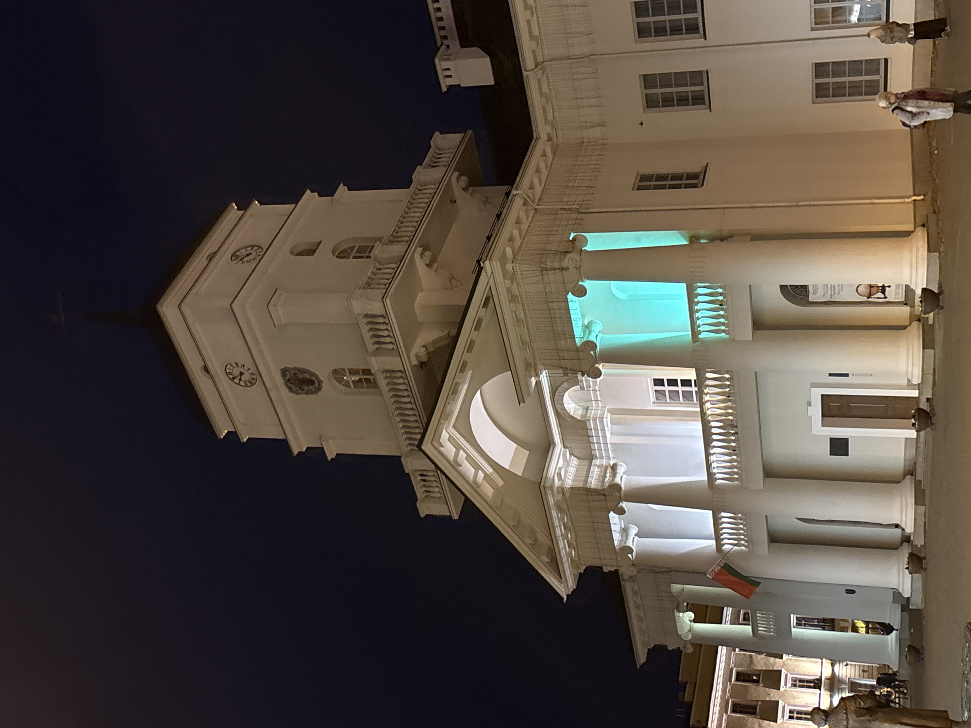 A low-angle view shows a tall, light-colored building with a clock tower at night, located in Minsk, Belarus. The building features classical architectural elements, including columns and ornate balconies. Some sections of the building are illuminated with a green light. A Lithuanian flag hangs from one of the columns. A few people are visible on the right side of the image. The sky is dark.