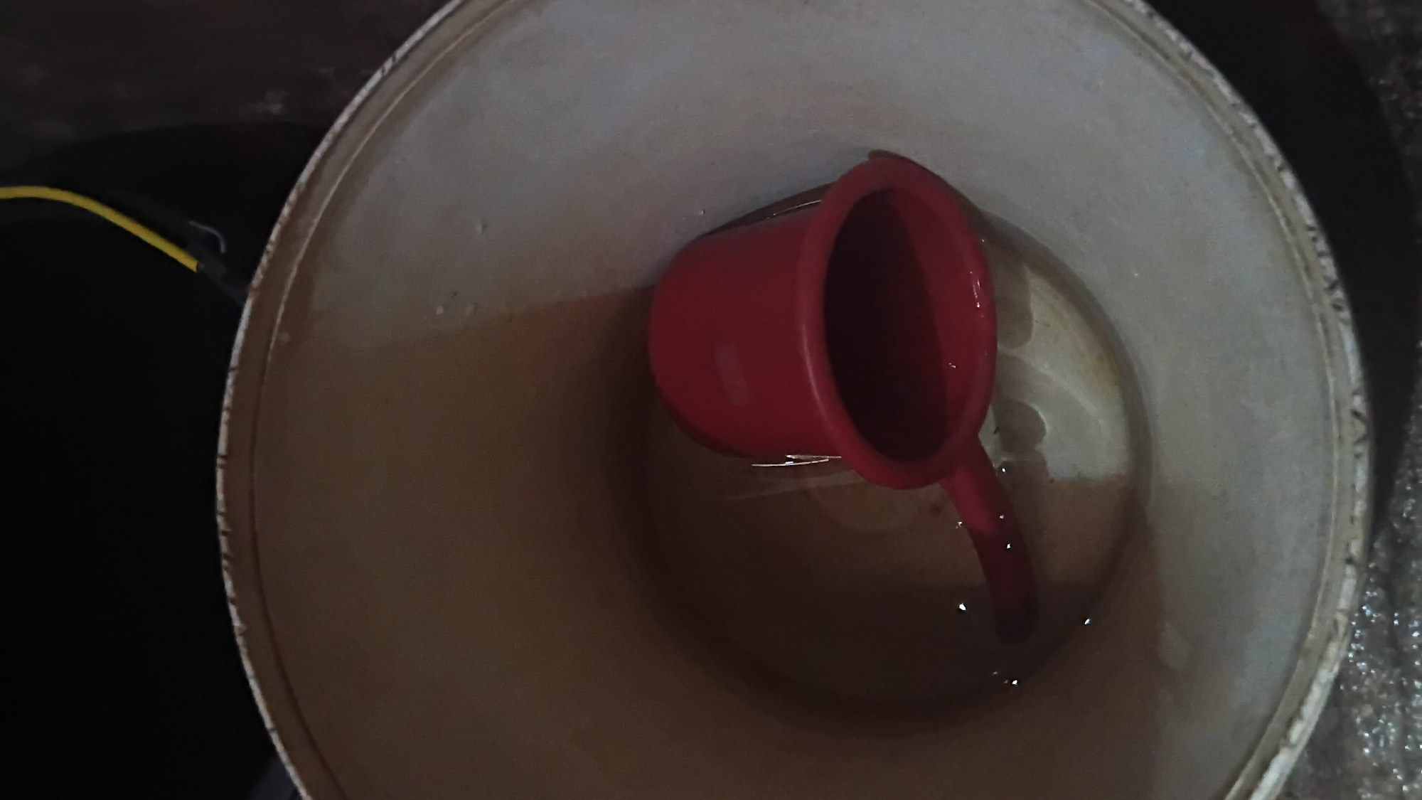 A red plastic dipper rests in a light brown, possibly beige, cylindrical container, which appears to be a bucket, containing a small amount of water. The water surface is disturbed, suggesting recent movement, and there are a few small droplets of water on the interior wall of the bucket above the water line. The dipper is positioned such that its open mouth is submerged in the water, and its handle extends outwards. The exterior of the bucket is not clearly visible, but the interior is textured and shows some signs of wear. The background is dark and out of focus, with a hint of a yellow wire visible at the top. The scene is indoors, with the floor beneath the bucket appearing wet and reflective, suggesting it may be tiled or a similar material. This image was captured within a residence in Prabumulih, South Sumatra, Indonesia.