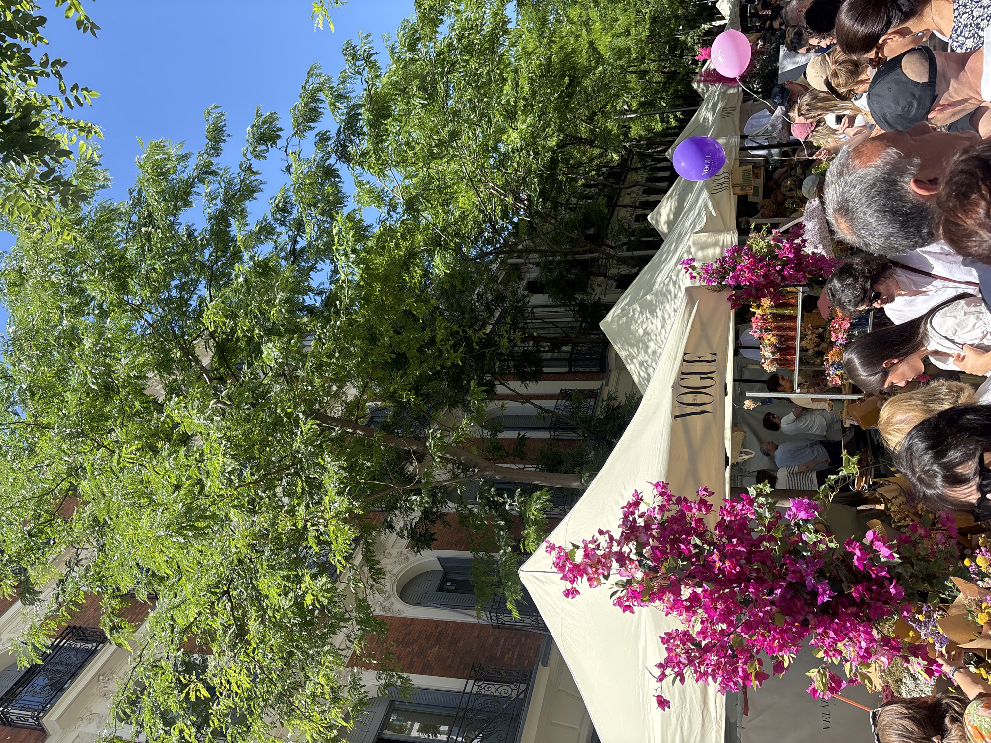 A street scene in Madrid, Spain, features several white canopies adorned with the word "VOGUE," likely indicating a fashion-related event. A crowd of people gathers around the canopies, suggesting commercial activity or an exhibition. Bright pink bougainvillea flowers are prominently displayed near one of the canopies, adding a touch of color. Two balloons, one pink and one purple, float above the crowd. Lush green trees frame the scene, providing a natural backdrop. In the background, buildings with brick facades and wrought iron balconies are visible, characteristic of Madrid's architecture. The sky is clear and blue.