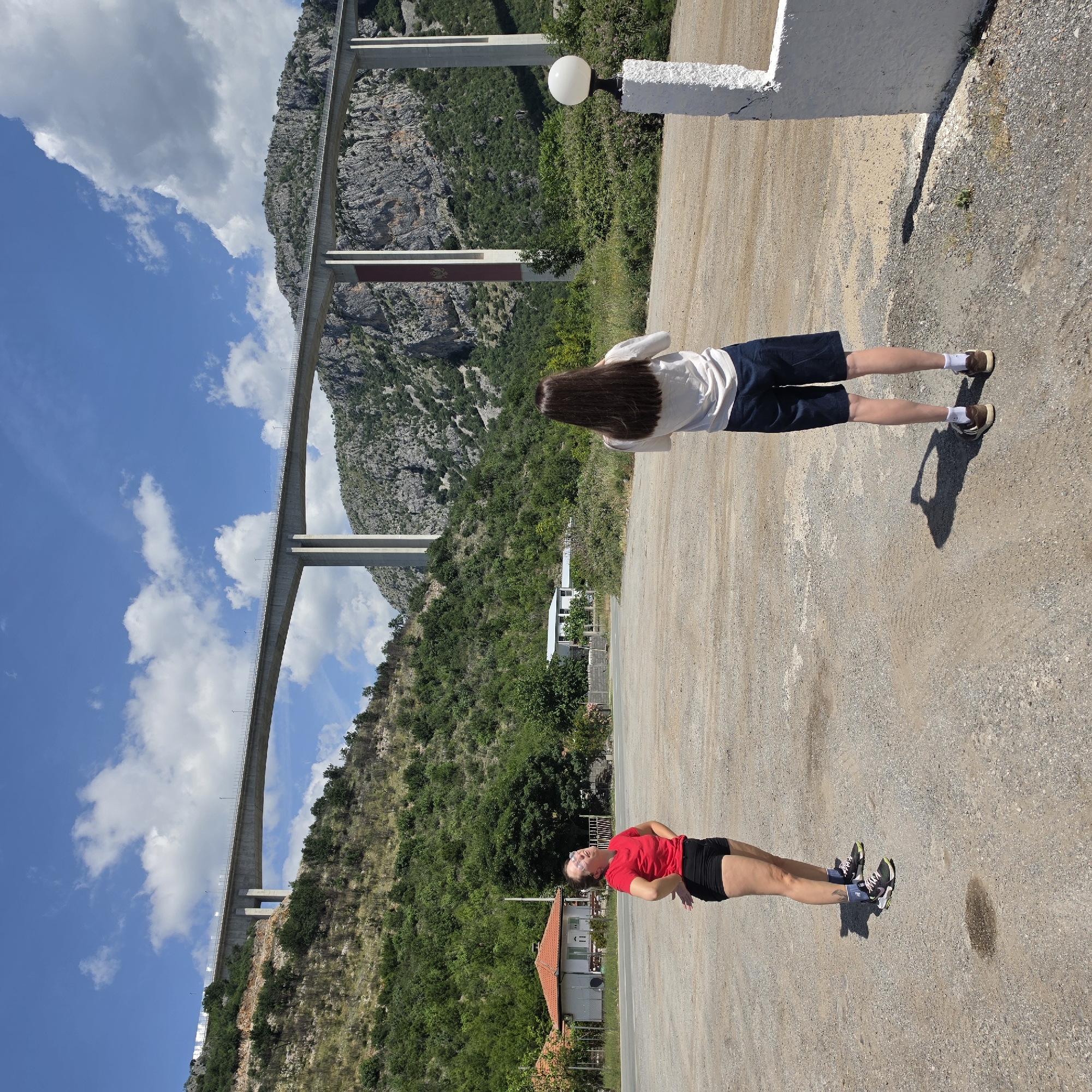 Two people stand on a gravel surface next to a white structure with a light fixture. In the background is the Moračica Bridge in Bioče, Montenegro, spanning a valley with green trees and gray rocks. One person wears a white shirt and navy shorts, facing away from the camera. The other person wears a red shirt and black shorts, facing the camera.