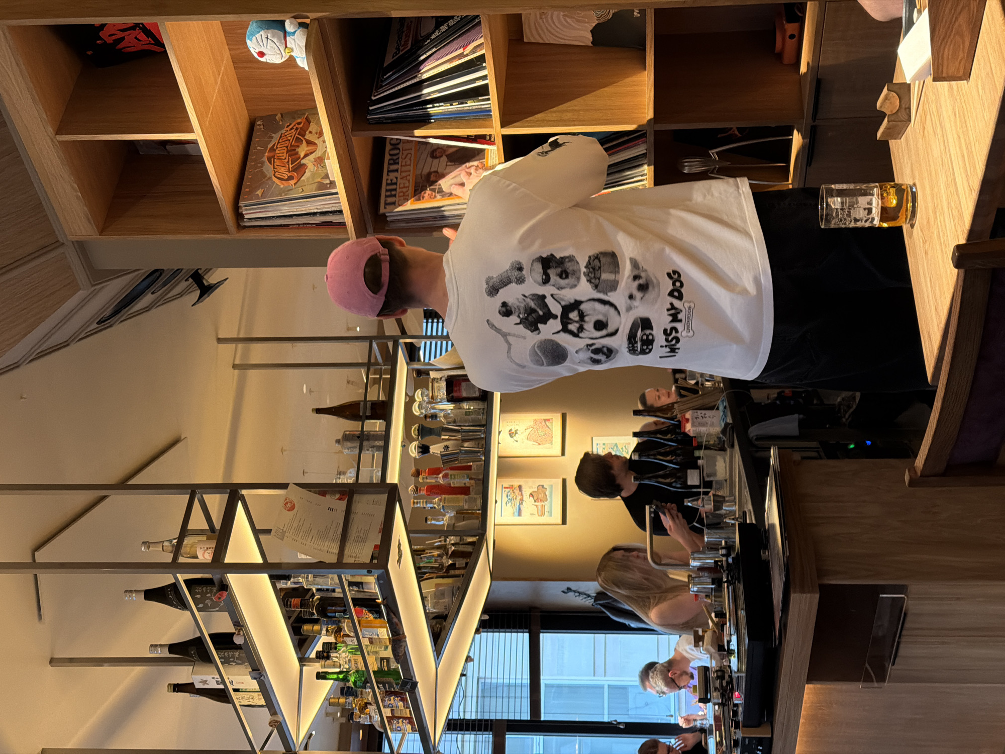 The image captures an indoor scene, likely a bar or restaurant, in Moscow, Russia, during what appears to be late afternoon or early evening, indicated by a mix of natural light from a window and warm artificial lighting. The perspective is looking towards the back of the establishment.
In the foreground, with their back to the camera, is a person wearing a white t-shirt, black pants, and a pink baseball cap. The t-shirt features a graphic design depicting several dog faces and dog-related objects like a bone and a tennis ball, accompanied by the text "MISS MY DOG" and a smaller "UNDERCOVER". Next to this person, on a wooden table, is a glass filled with an amber-colored liquid, likely an alcoholic beverage.
Behind the person, a tall wooden shelving unit is filled with various items, including books, magazines, and what appear to be vinyl records. A blue Doraemon plush toy is visible on one of the shelves.
Further into the background, a bustling bar area is visible. Two individuals are behind a long wooden bar counter: one with dark hair looking downwards, and another with blonde hair, partially obscured. They appear to be tending to the bar or engaged in conversation. Above the bar counter, a striking, multi-tiered metal shelving unit, illuminated from below, holds a wide array of bottles, including spirits, wine, and several sake bottles with Japanese script. Some bar tools like shakers are also visible.
Framed artwork hangs on the wall behind the bar. To the left, a large window with horizontal blinds offers a glimpse of the exterior, showing parts of a building. The overall ambiance is social and relaxed, consistent with a contemporary bar environment.