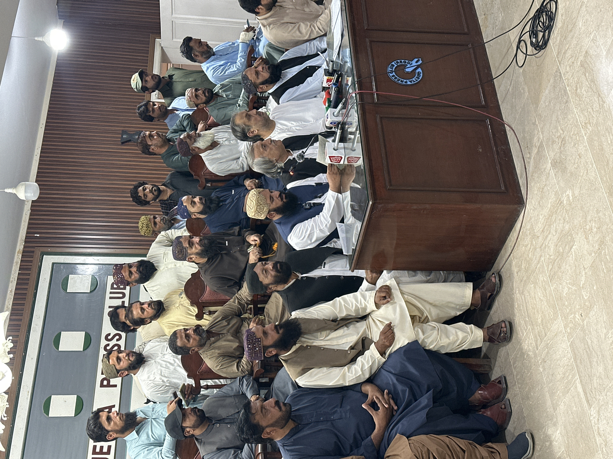 The image shows a group of men seated indoors, likely at a press conference. They are gathered behind a table with microphones and a logo indicating "Quetta Press Club." The men are dressed in various traditional Pakistani clothing. The location is in Quetta City Tehsil, Pakistan, based on the logo of the Quetta Press Club on the table.