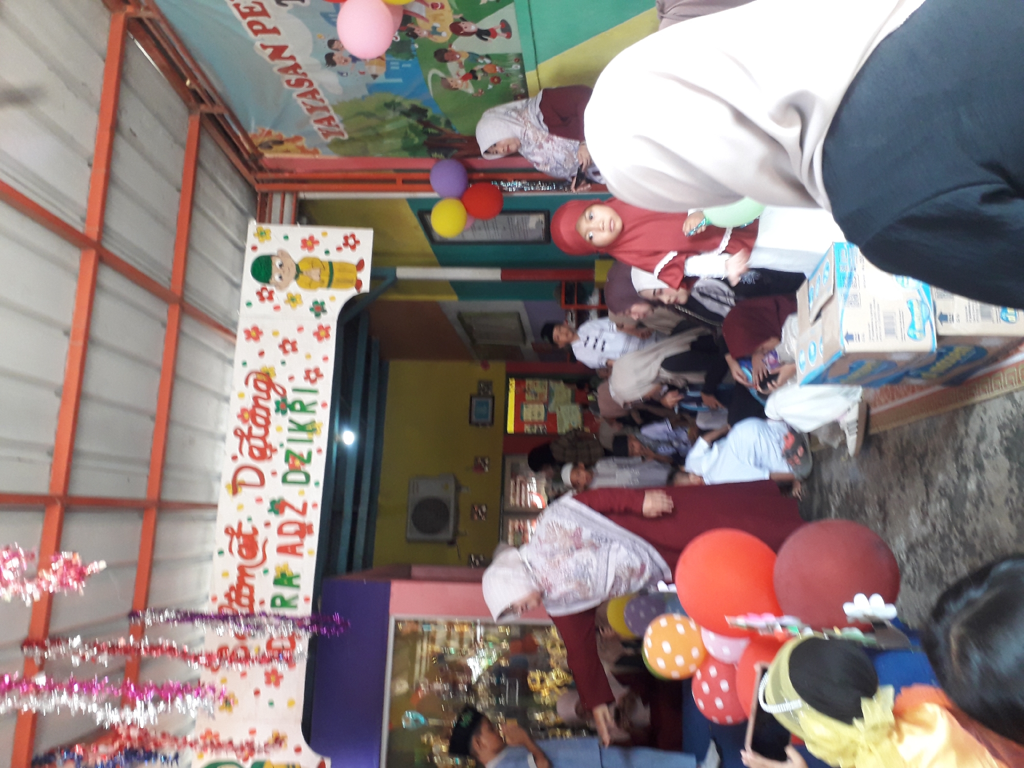 The image depicts an indoor gathering in Cankuang, Indonesia, likely a school or community event. The environment appears festive, with colorful streamers hanging from the ceiling and balloons scattered around. In the background, a banner with Indonesian text, possibly a greeting or event title, is visible. Several people, including adults and children, are present, many wearing traditional Islamic attire like hijabs and peci caps. Some individuals are standing, while others are seated. Children are interacting with balloons, and there's a general atmosphere of celebration. The presence of a display case with trophies in the background suggests an educational institution.