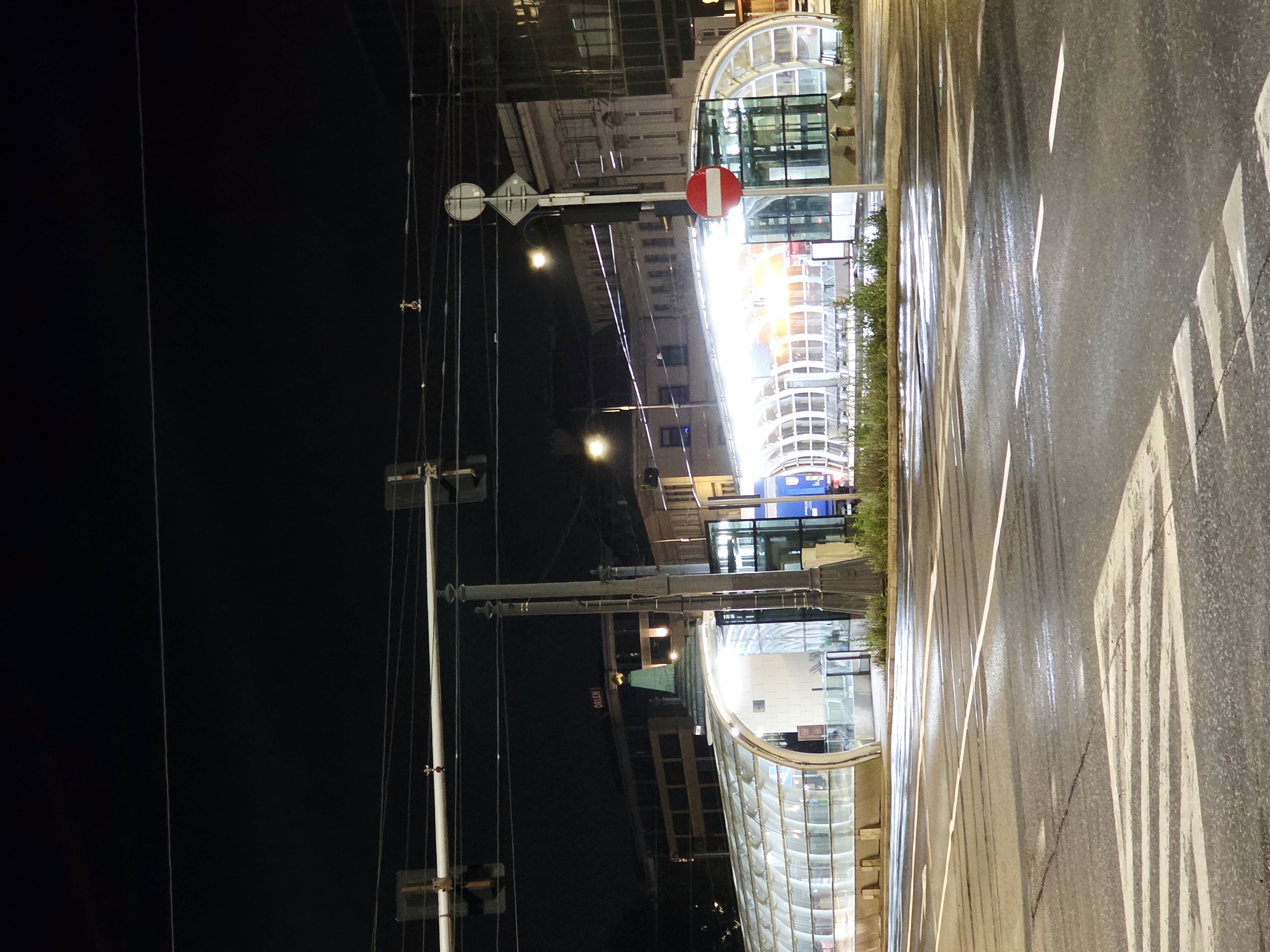 This is a nighttime photograph of a street scene in Warsaw, Poland, likely near the Centrum metro station. The foreground shows a wet asphalt road with white lane markings and a crosswalk. To the right, a modern, curved glass and metal structure that appears to be an entrance to an underground facility or public transportation station is illuminated. It has a sleek, contemporary design.
Behind the glass structure, various buildings are visible. One building has a sign that reads "ORLEN." Another building to the right has a classical architectural style with columns and arched windows, reflecting the city's varied architectural heritage. Overhead, traffic lights and power lines crisscross the dark sky. A red and white circular traffic sign indicating "no entry" is mounted on a pole to the right of the glass structure. The overall atmosphere is urban and somewhat sterile due to the nighttime lighting and the wet road surface.