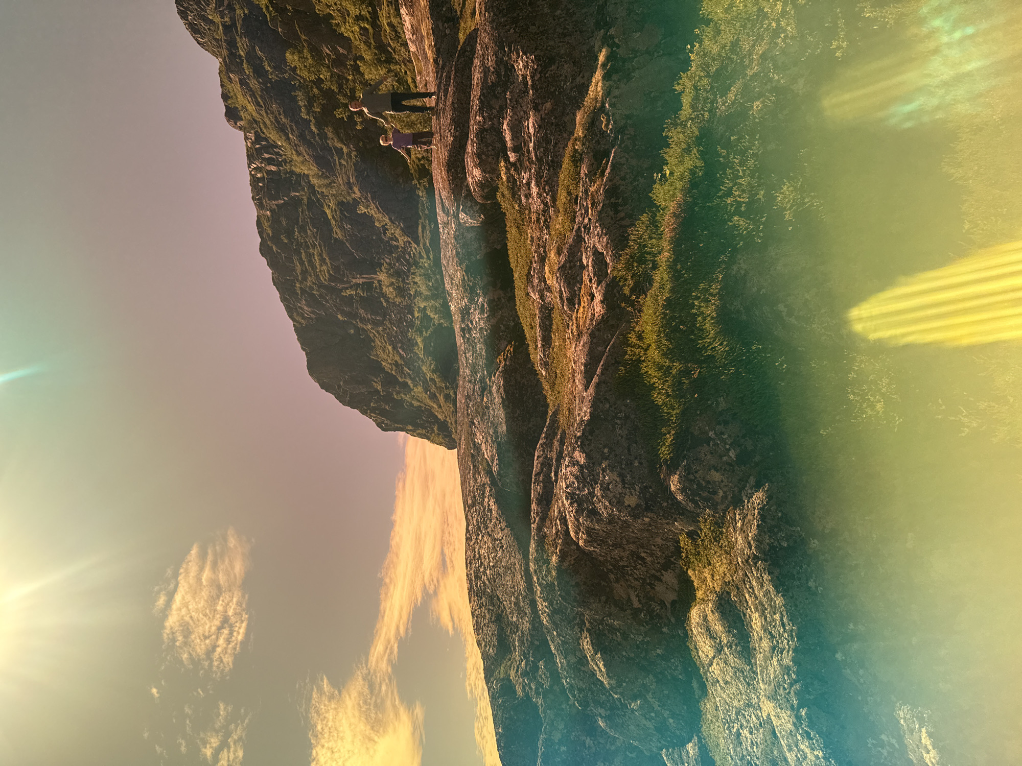 A man and a child stand on a rocky, grassy hillside under a bright sun with lens flare. The man, wearing a grey shirt and dark pants, stands on a higher rock formation, looking towards the child. The child, wearing a purple shirt and brown shorts, stands on a lower rock formation, facing forward with arms outstretched. The hillside is covered in rocks and vegetation, with a steep cliff face rising behind them. The sky is a light blue with streaks of yellow and pink clouds. The image appears to have a vintage or artistic filter applied, with colored light leaks visible at the bottom and top edges. The scene is set in Teriberka, Russia.