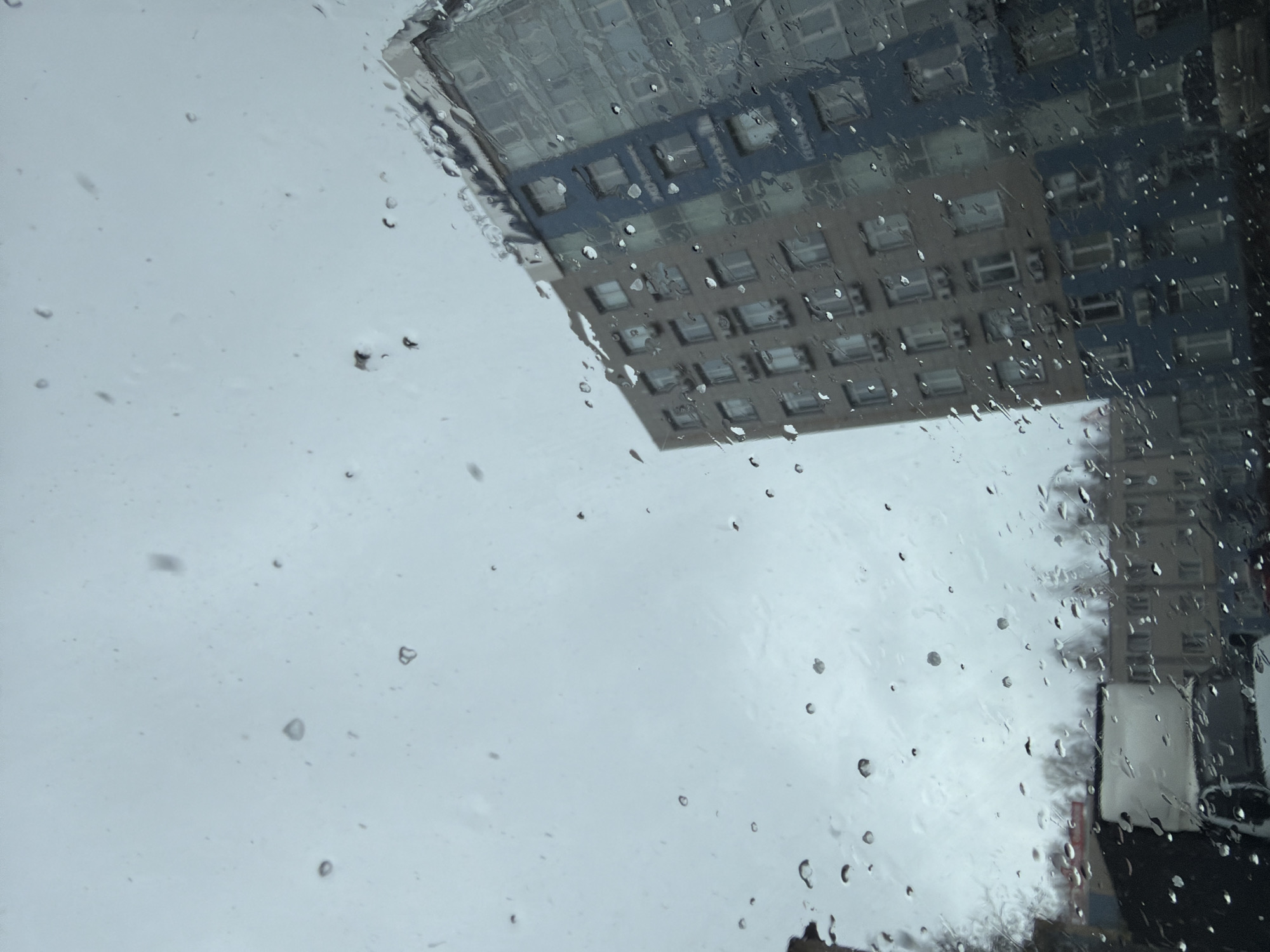 The image depicts a view through a wet window, possibly a car window due to the distorted reflection of buildings. Raindrops cover the glass. The dominant features are two high-rise buildings reflected in the window, with a grey sky visible behind them. The building on the left is multi-story with a grid-like pattern of windows. The building on the right is obscured by raindrops and other objects. The location is likely in the city of Chaikovsky, Russia. There are no people visible. The scene suggests rainy weather.