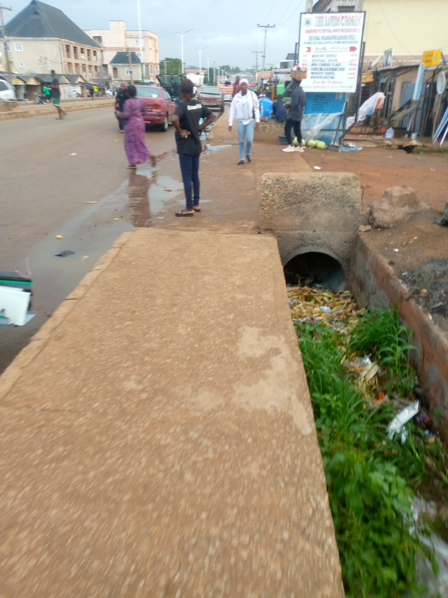 The image depicts a street scene in Bukuru, Nigeria. A concrete drainage ditch runs along the foreground of the image, filled with trash and vegetation. Several people are visible along the street. To the left, a woman in a purple patterned dress is walking. Next to her, a person wearing a black shirt and jeans stands near the drainage ditch. Further down the street, a person in a white jacket and jeans walks toward the camera. To the right of them, a man in a hat and dark clothing stands next to a sign that reads "THE LORD'S CHOSEN". The street is wet. Cars are visible in the background. Buildings are situated on both sides of the street. The sky is overcast.
