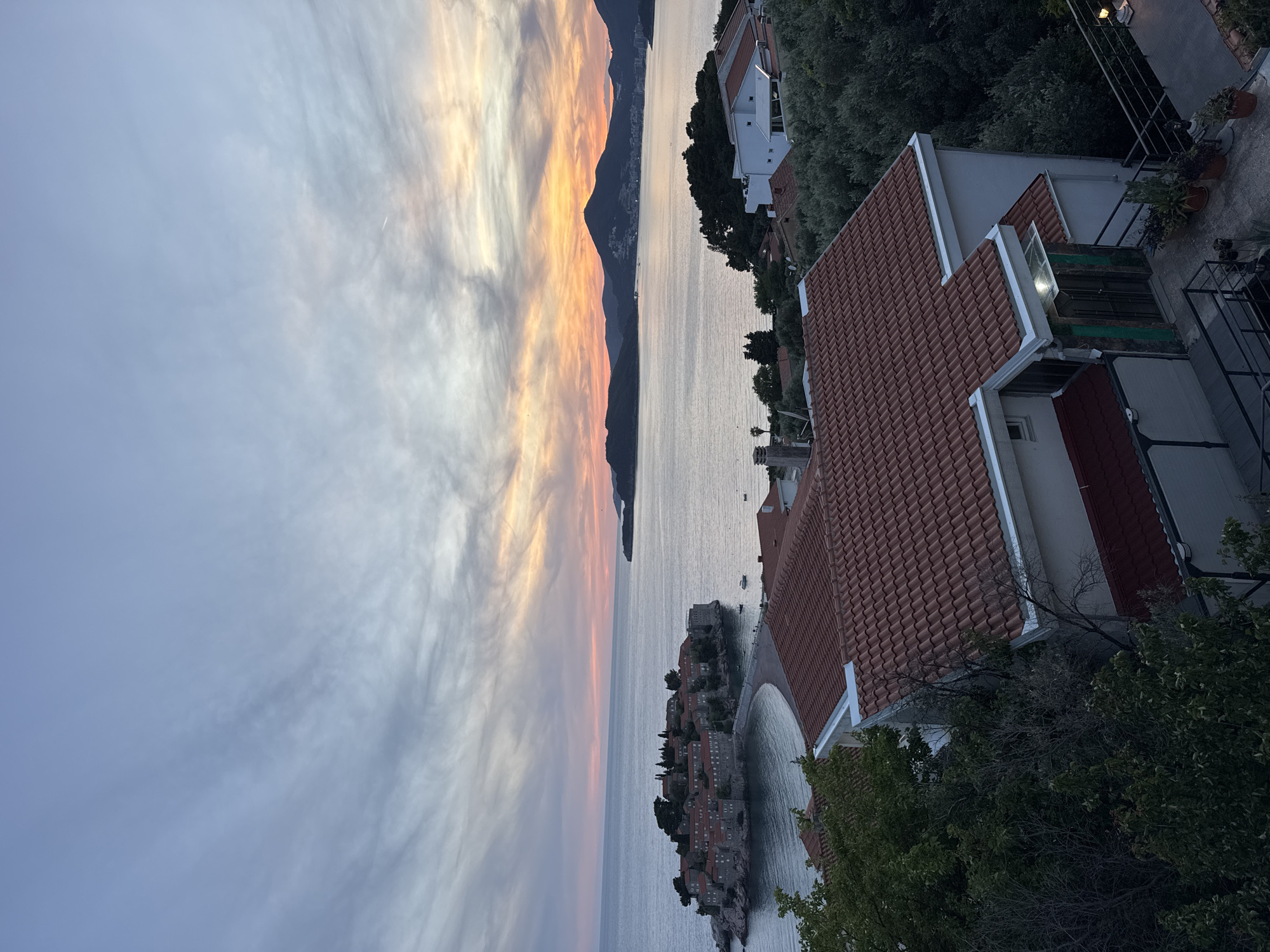 The image captures an elevated view in Sveti Stefan, Montenegro, showcasing a picturesque coastal scene at what appears to be dusk or dawn. A dramatic sky with orange and gray-streaked clouds fills the left side of the frame. The Adriatic Sea stretches horizontally across the center of the image, reflecting the sky's colors. A dark, mountainous landmass is visible in the distance.
In the lower-center, a small, densely built island with red-roofed buildings, characteristic of Sveti Stefan, is connected to the mainland. The island's architecture is traditional, with closely packed structures.
On the right side of the image, the tiled roof and balconies of a building are visible, suggesting the perspective is from a higher vantage point within the town. Green trees and foliage frame the building, partially obscuring the view.
The overall impression is of a serene, natural landscape combined with the architectural charm of a historic coastal town.