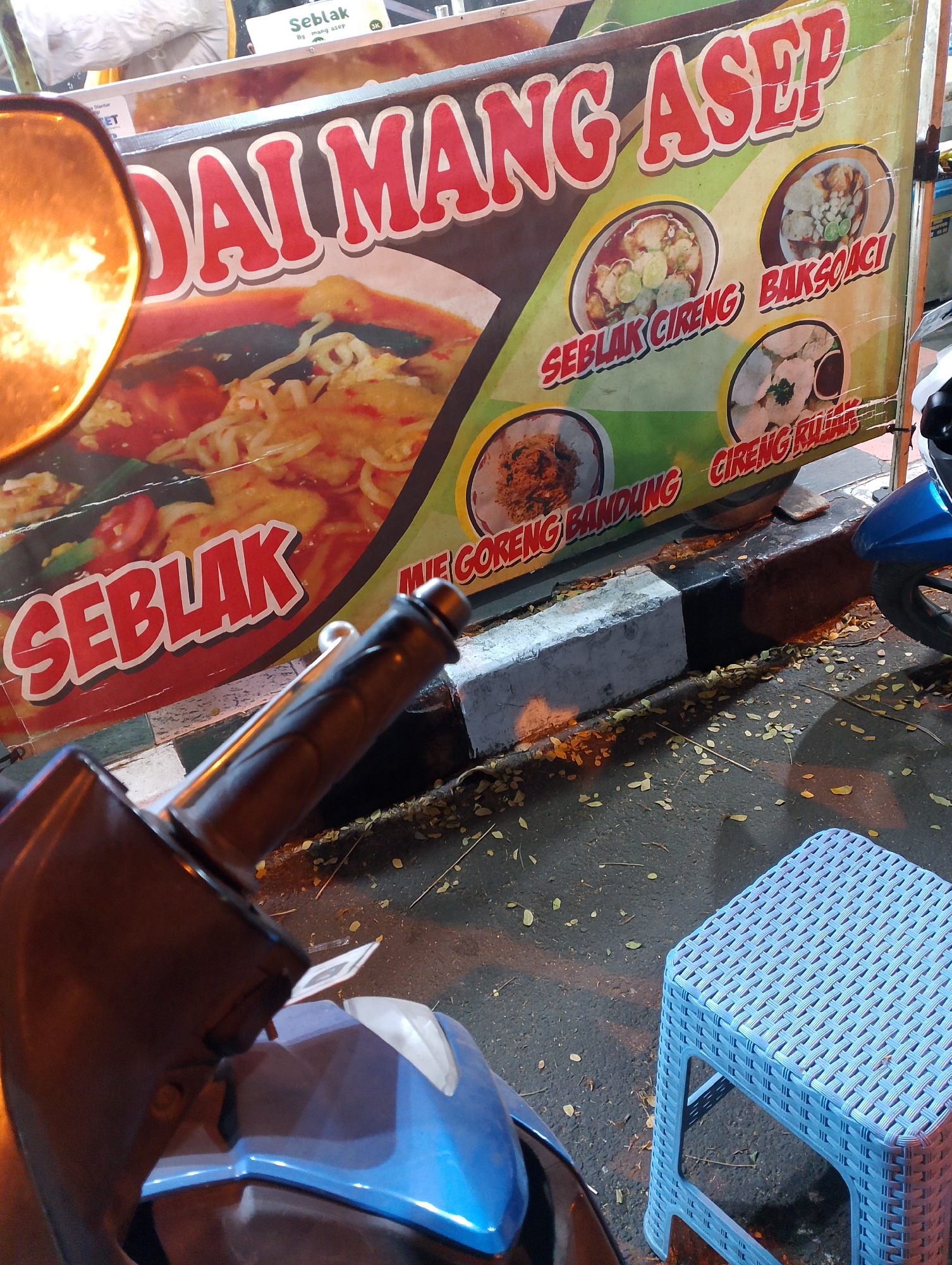 A banner advertising a food stall is displayed horizontally. The banner prominently features the words "DAI MANG ASEP" in large red lettering. Below this, the word "SEBLAK" is written in red on the left side. The banner also shows images of various dishes, including "SEBLAK CIRENG," "BAKSO ACI," "MIE GORENG BANDUNG," and "CIRENG RUJAK." The stall appears to be outdoors, on a street with fallen leaves on the ground. A blue plastic stool is visible in the foreground, and part of a blue scooter is also present. The lighting suggests it is nighttime. The banner includes a small sign with the word "Seblak" and "BK" which could indicate the location of the stall as Magelang, Indonesia, given the common use of abbreviations and local street food advertising styles in that region.