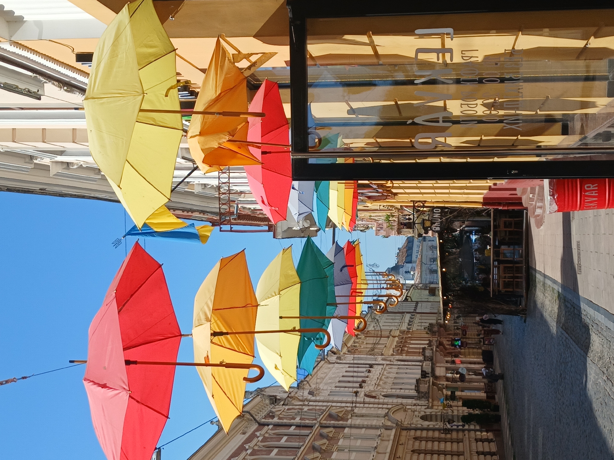 An eye-level, medium shot captures an outdoor scene on a cobblestone street lined with multi-story buildings. The sky is clear and blue. Overhead, a series of colorful umbrellas are suspended, creating a vibrant canopy. The umbrellas are arranged in rows, with red, yellow, orange, green, blue, grey, and white varieties visible. They hang by thin wires, and their handles are visible.
On the right side of the frame, a building facade is painted in a warm yellow hue. A large glass window with a black frame is prominent, reflecting the yellow wall and casting shadows. White lettering is visible on the glass, appearing reversed. A red cylindrical trash can with white lettering reading "EKVÁR" is positioned on the sidewalk to the left of the window.
The street itself is paved with cobblestones. On the left side of the street, the buildings exhibit classical architectural details, with arched windows and decorative trim. Some smaller signs and lights are visible on these buildings. A few people are present on the street, appearing as small figures in the distance, walking and interacting.
Further down the street, a covered outdoor seating area with tables and chairs suggests a cafe or restaurant. In the background, more buildings are visible, with a church or similar structure with a dome shape in the far distance. Two Ukrainian flags, one blue and one yellow, are also visible hanging from a building on the right side of the image. The overall impression is a lively, decorated urban street.