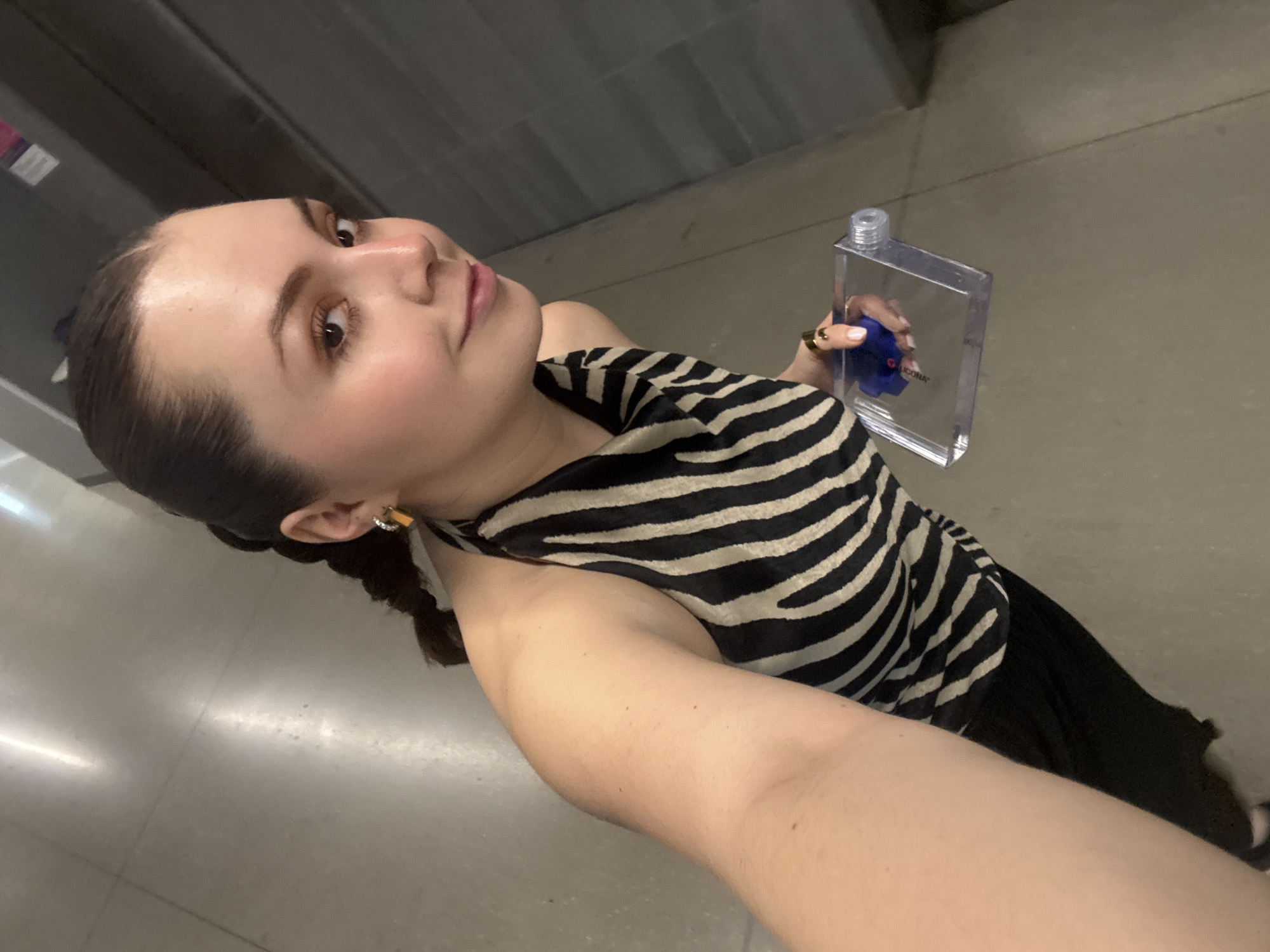 A woman is holding a clear, rectangular water bottle with a blue interior component. She is wearing a black and white striped halter top. She also has a gold ring on her finger and a gold square earring. The environment appears to be an interior space with concrete floors and walls. It is undetermined if it's Mexico City, Mexico.