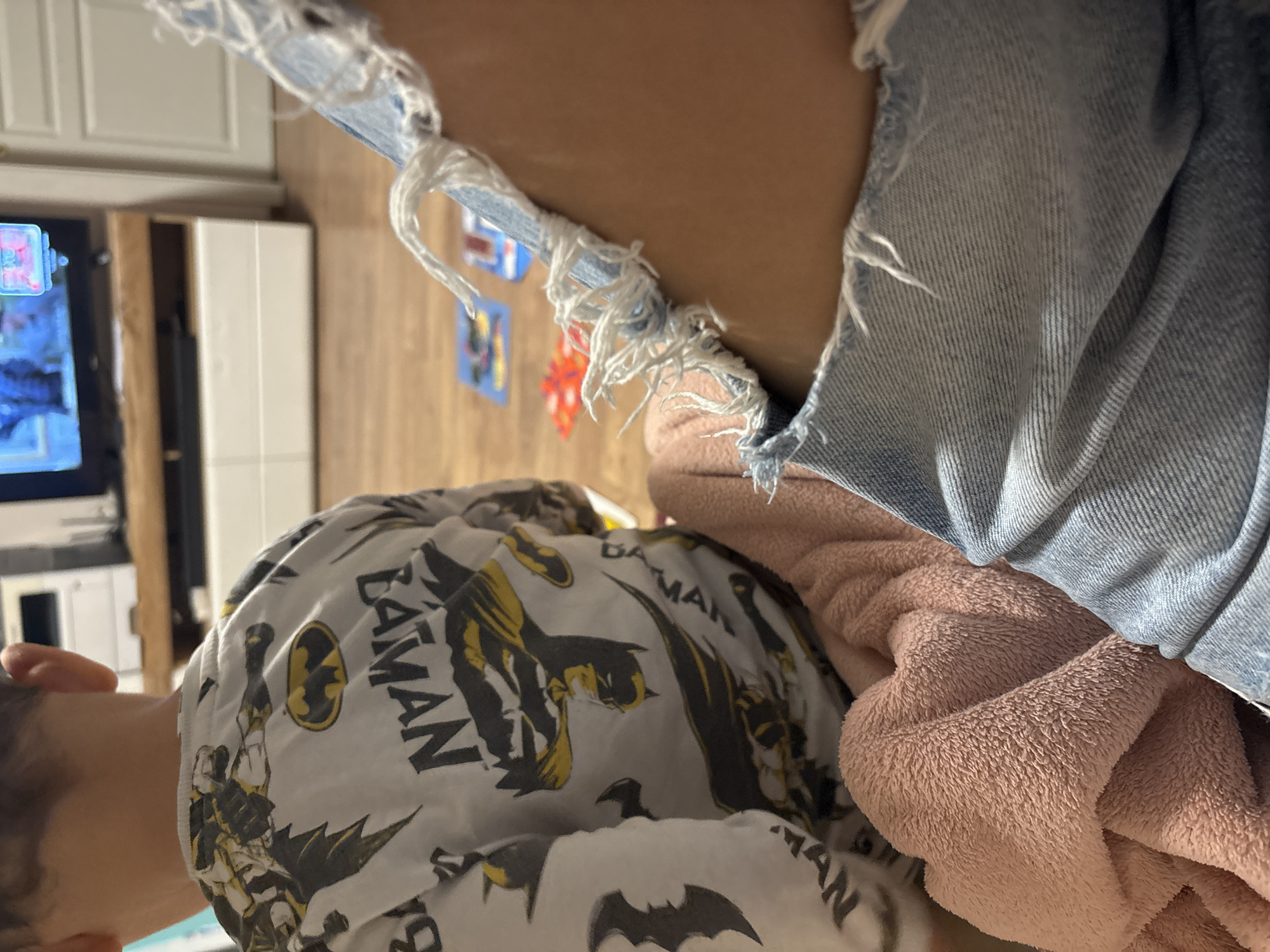 The image, captured in Plovdiv, Bulgaria, depicts an indoor domestic scene with a rotated perspective. In the lower left, partially visible from the back and side, is a person, likely a child, wearing a white short-sleeved shirt patterned with various black and yellow Batman designs. The shirt features the Batman logo and multiple illustrations of the superhero, along with the visible text "BATMAN" repeated across the fabric.
In the upper right, another person's leg and hip area are visible, clad in light blue, distressed denim shorts or jeans. These garments feature prominent rips and frayed edges, revealing skin underneath. A soft, pink, fuzzy blanket is draped beside this person, appearing warm and comfortable.
The background shows elements of a home environment. A television screen, partially visible on the left, displays bright, colorful content that appears to be a video game or an animated program. Adjacent to the television, there is a white cabinet and light-colored wooden wall paneling or flooring. Several small, colorful pictures or decorative items are affixed to the wooden surface. The lighting suggests it is daytime. The overall impression is one of casual relaxation within a living space.