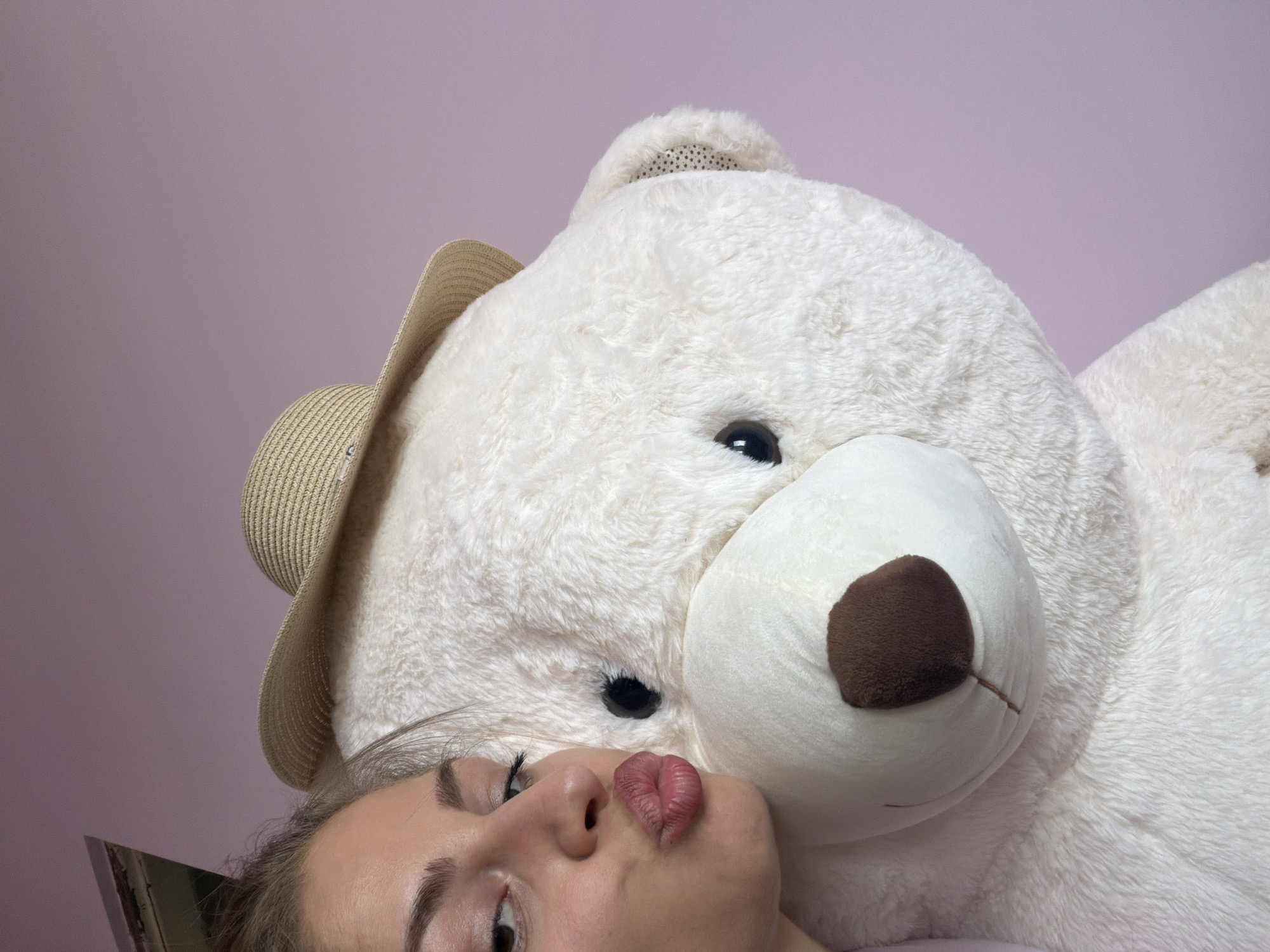 The image shows a close-up of a woman and a very large, white stuffed teddy bear. The woman is on the left side of the frame, leaning in and puckering her lips towards the bear's face. She has dark hair pulled back and is looking towards the camera. The teddy bear is wearing a straw hat and is positioned to the right of the woman, filling a significant portion of the frame. The background is a solid light purple wall. There are no identifiable landmarks or features that would indicate the specific location within Etropole, Bulgaria. The scene appears to be indoors.