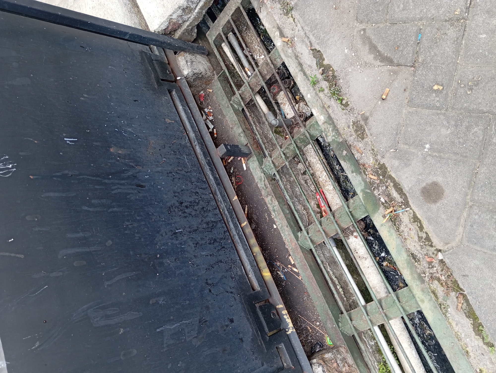 The image shows a close-up, eye-level view of a metal grate over a drainage ditch. The grate is old and rusty, with green paint peeling off in places. Beneath the grate, the ditch is filled with dark, murky water and debris, including cigarette butts and what appears to be trash. Several pipes run through the ditch, some of which are white and some are gray. The ground around the grate is paved with gray cobblestones, and there is a large, dark metal door or gate behind the grate, which is also dirty and worn. The scene appears to be located in an outdoor urban environment, possibly along a street or sidewalk in Nogosari, Indonesia, given the context provided, as suggested by the presence of cigarette butts which are common litter in many public spaces. The overall impression is one of neglect and disrepair.