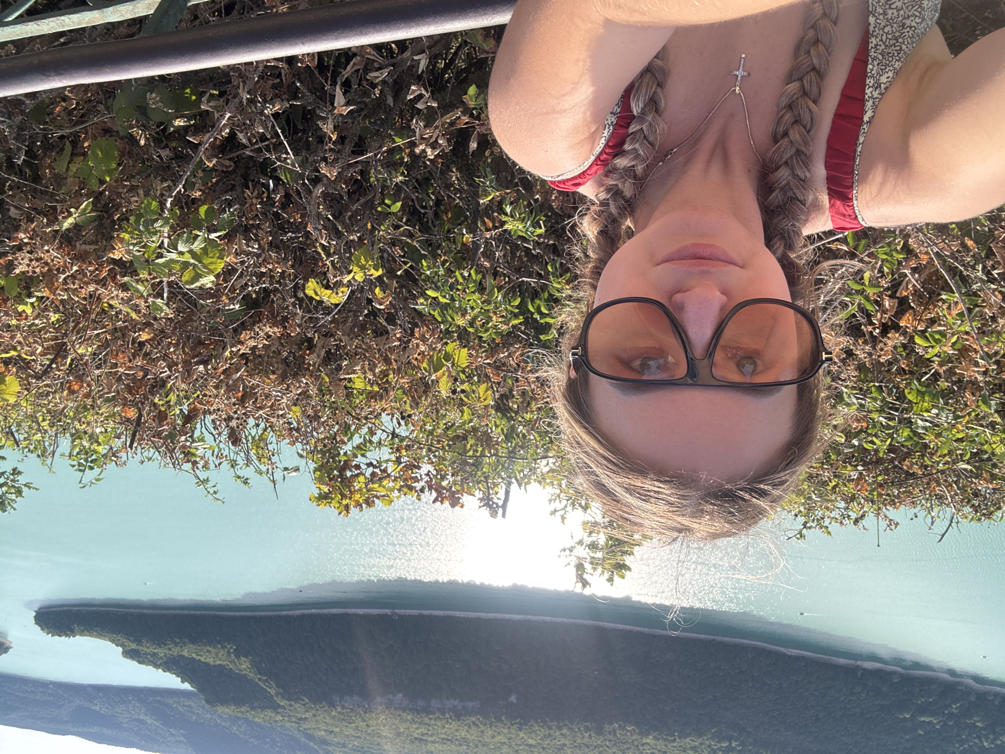 A young woman is taking a selfie outdoors with a body of water and forested hills in the background. The woman is wearing large, dark-rimmed sunglasses with orange lenses. Her blonde hair is styled in two braids. She is also wearing a red and white patterned top and a silver necklace with a cross pendant. The woman is positioned in the foreground, with a dense thicket of brown and green foliage to her right. Behind the foliage, a wide expanse of turquoise water is visible, reflecting the sunlight. The water is bordered by lush, green, tree-covered hills, characteristic of the region surrounding Moirans-en-Montagne, France. The sky above is clear and bright. A dark metal railing is visible along the bottom of the frame.