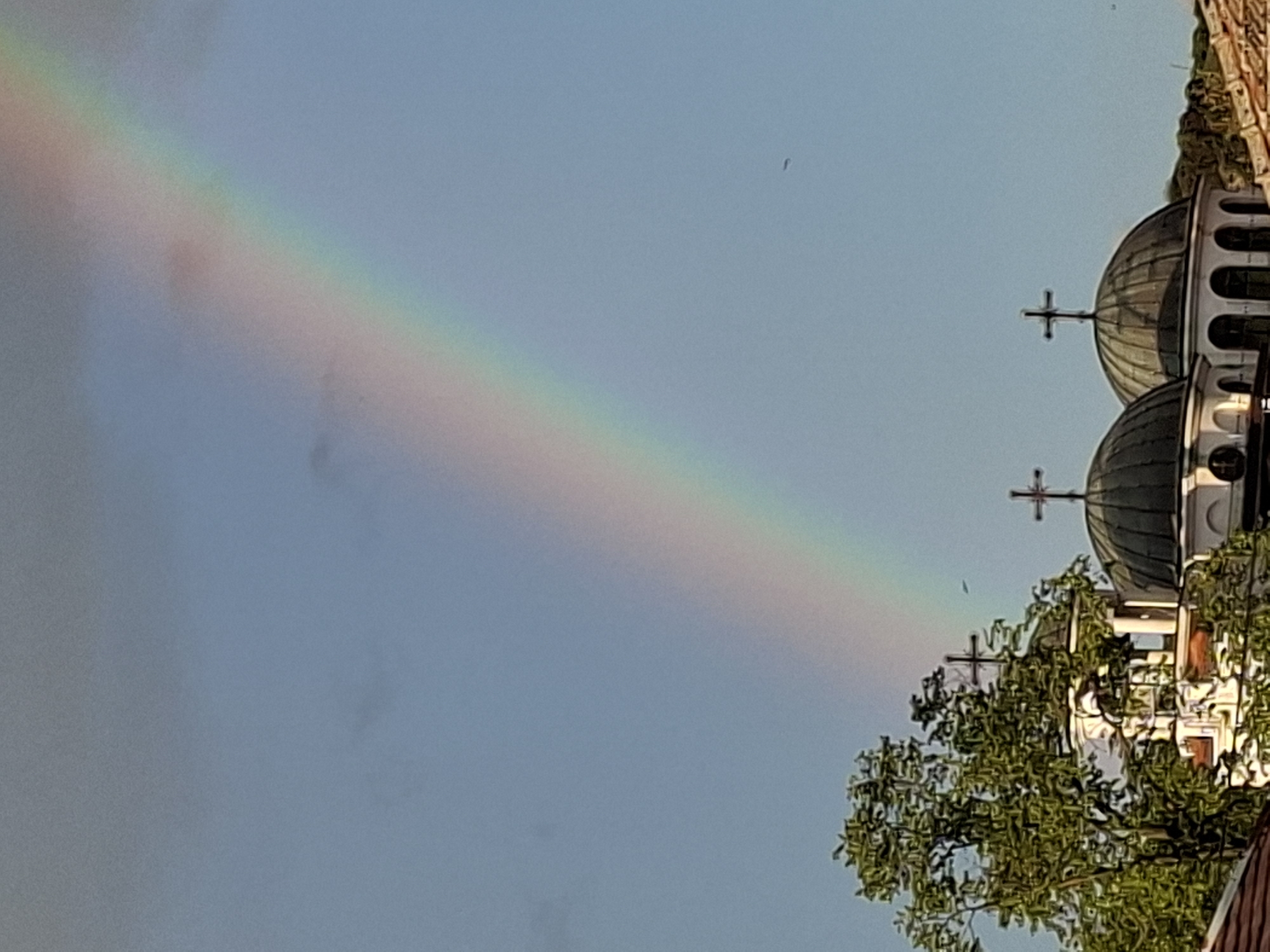 The image shows a rainbow spanning across a blue sky. The rainbow arches over a building with domes topped with crosses. A tree with green leaves is in the foreground near the building.