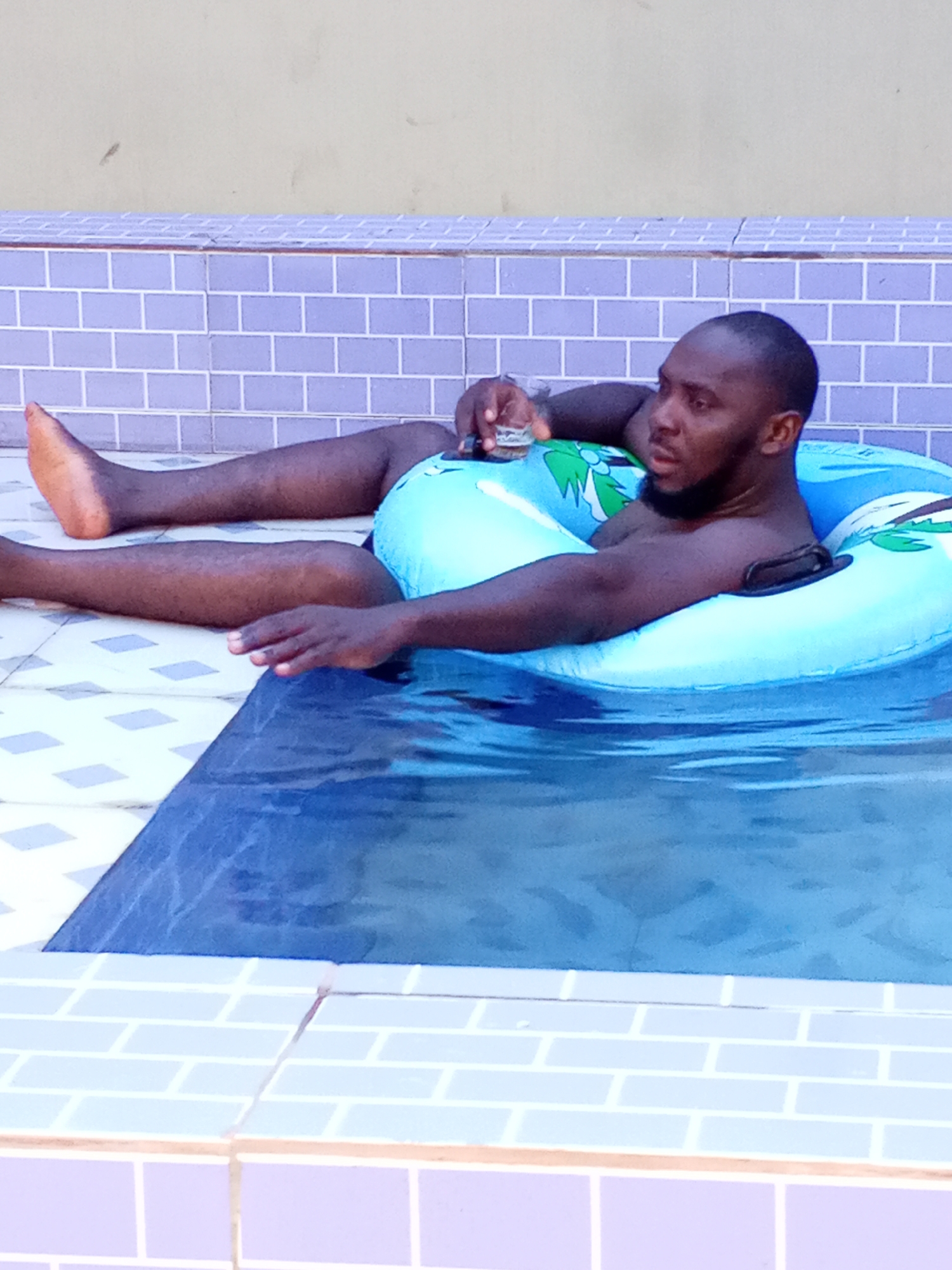 A man is relaxing in a blue inflatable ring in a swimming pool. He is holding a small glass in his left hand. The pool is bordered with light blue and purple tiles. His legs are extended on the tiled patio. The location is Ankpa, Nigeria.