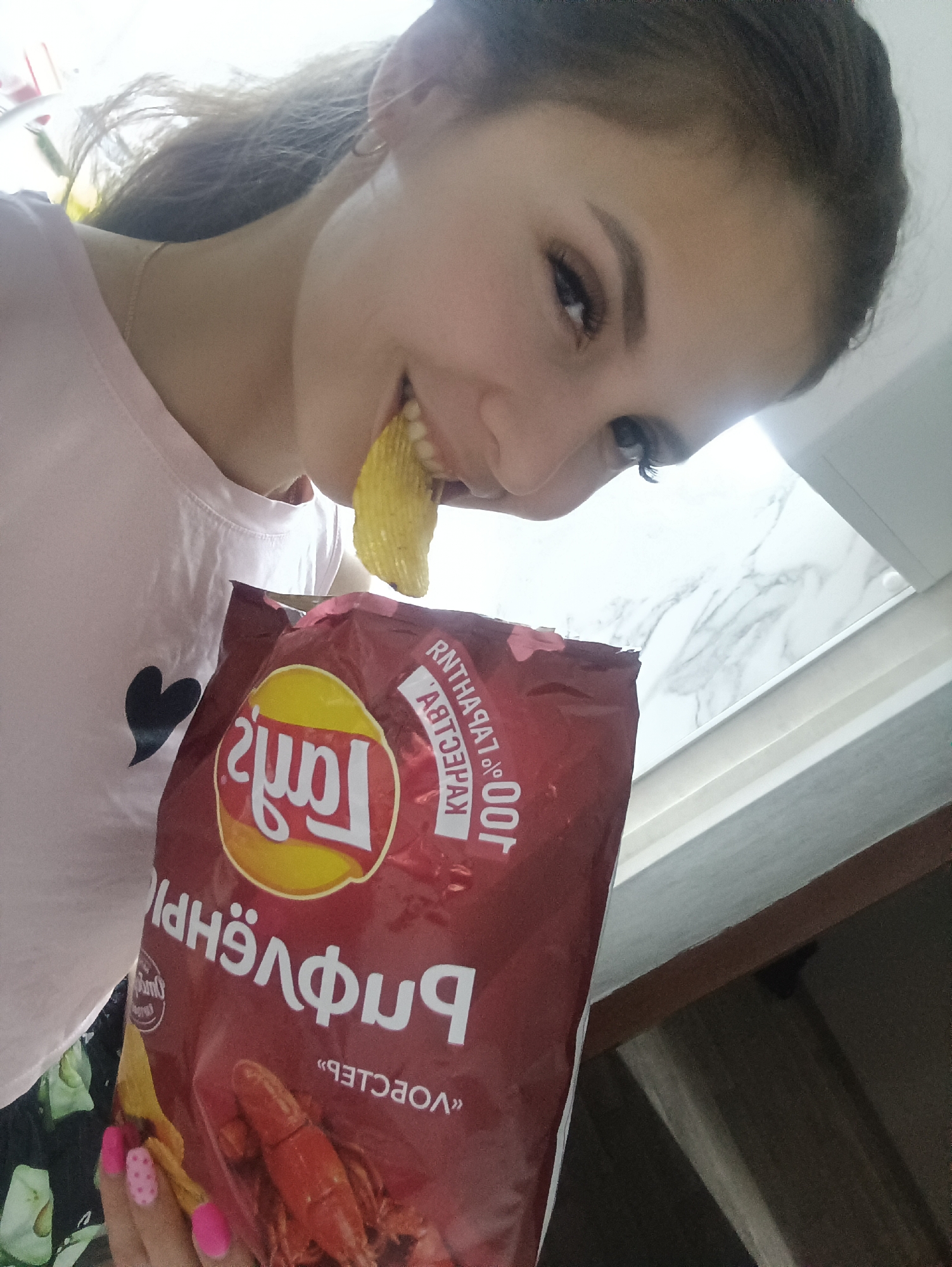 A young woman is pictured from a high angle, holding a bag of Lay's "Crayfish" flavored potato chips and eating one. She has light brown hair, light skin, and is wearing a light pink t-shirt with a black heart on it. Her nails are painted pink, some with white polka dots. The background is blurred, showing a white marble-patterned surface and a wooden cabinet. The text on the chip bag is written in Russian. The location appears to be indoors, possibly a kitchen.