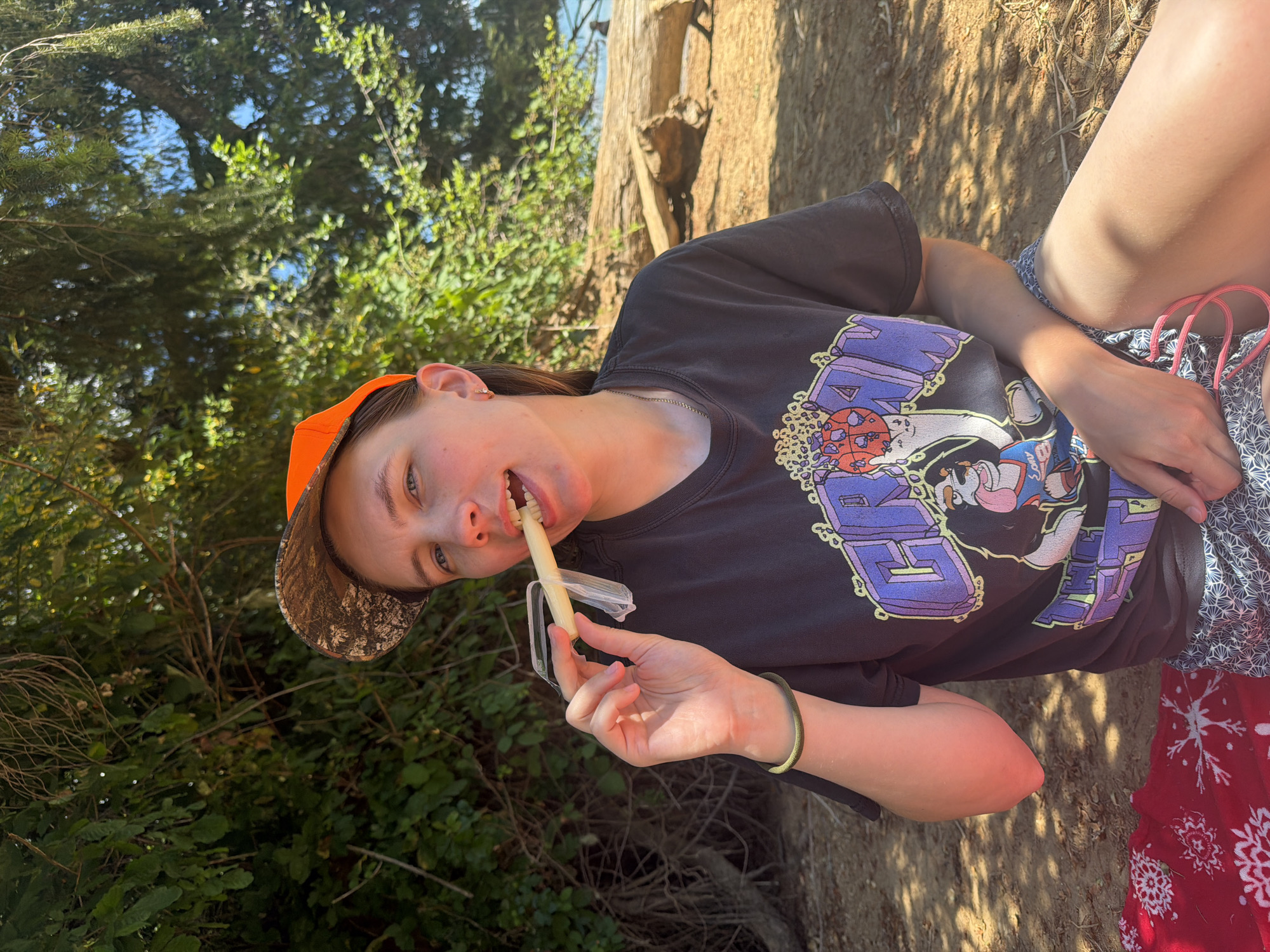 A young adult wearing an orange and camouflage baseball cap, a black t-shirt with a cartoon graphic, and patterned shorts sits outdoors on a sandy area. They are holding a white, cylindrical object, possibly food, to their mouth with their right hand, which is also wearing a green bracelet. The background features lush green trees and a large fallen log. Sunlight filters through the foliage, casting shadows on the ground. The overall setting appears to be a natural, possibly coastal, environment. The location is identified as Cherry Grove, United States.