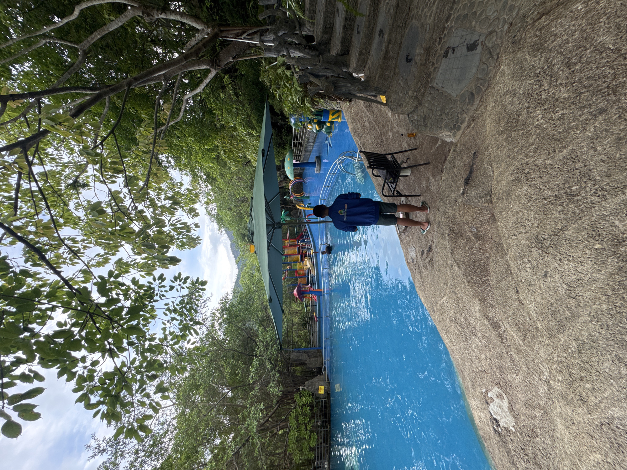 An outdoor scene captures a vibrant swimming pool area, likely a resort or park in Đà Nẵng, Vietnam. The image is taken from a slightly elevated perspective, looking down towards a bright blue swimming pool surrounded by lush green foliage and natural-looking rock formations.
The main subject is a person, appearing to be a child or young teenager, standing with their back to the camera at the edge of the pool. They are wearing a blue long-sleeved top or jacket, dark shorts, and sandals, with their lower legs either in or very close to the water. In the pool, partially obscured by a large green shade umbrella, is colorful play equipment, including a small slide and rings, with another person (possibly a child in pink swimwear) visible near these structures.
The ground surrounding the pool is textured and rocky, with some areas featuring patterned stone tiles. A black metal chair and a yellow-capped bottle rest on this ground. Dense trees with abundant green leaves create a natural canopy, with their branches stretching across the upper part of the frame, and more foliage forming a thick background. The sky is visible through the tree cover, showing bright white clouds, indicating daytime. Metal railings are present around the pool edge and leading up some stone steps. While a small yellow sign is visible on a railing, its text is unreadable. The scene conveys a relaxed, recreational atmosphere.