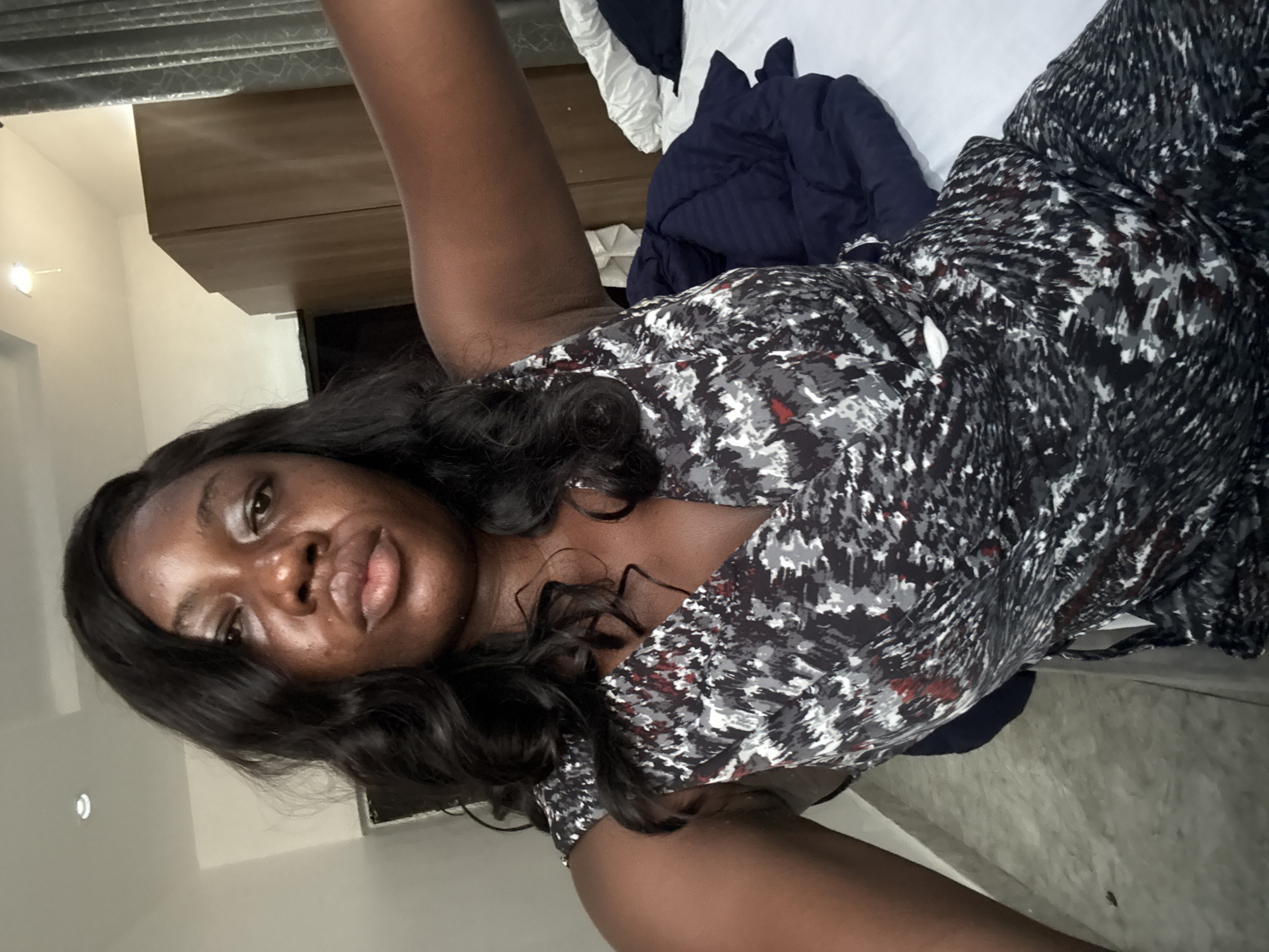 An indoor, medium shot captures a woman lying down, looking towards the camera, with the image rotated 90 degrees clockwise. She has dark skin and long, wavy dark hair. She wears a V-neck top or dress patterned with black, white, grey, and subtle red specks. Her expression is neutral.
Behind her, a bed is visible with white sheets and a dark blue blanket or duvet. To her left (viewer's top right), there is a piece of wooden furniture, possibly a headboard or a dresser. To her right (viewer's bottom left), a light-colored wall is visible with a small, white wall-mounted light fixture. The lighting appears to be artificial and internal, suggesting an indoor setting. No text is visible in the image. The setting appears to be a bedroom or hotel room in Iddo Tudunwada, Nigeria.