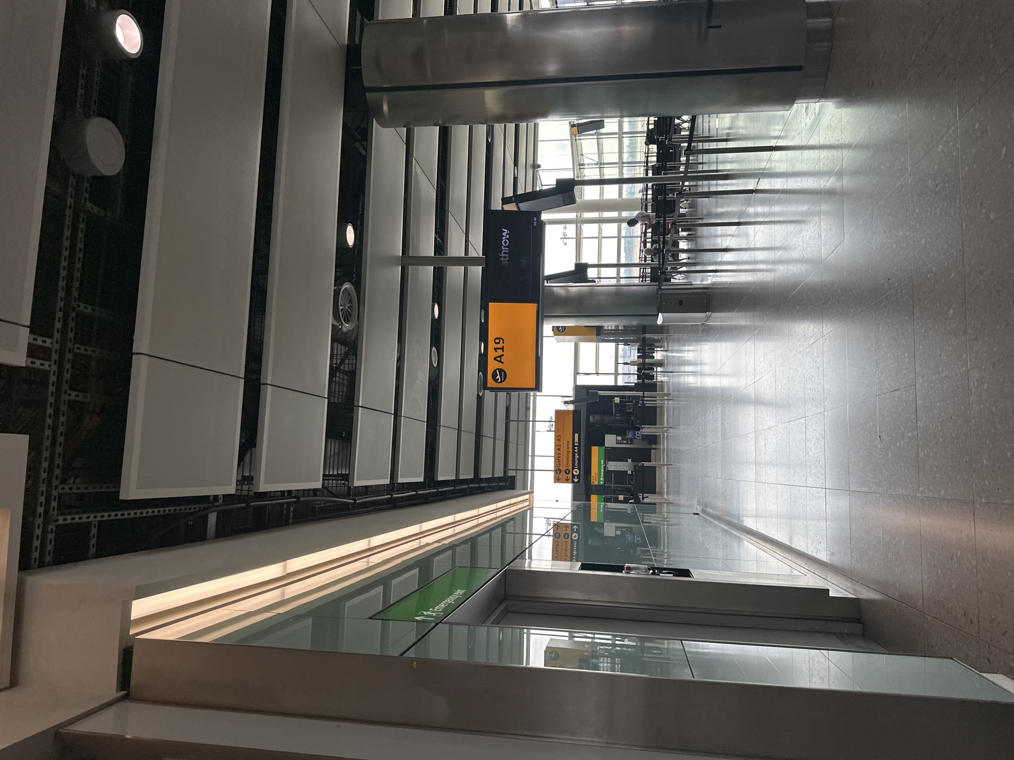 This image depicts an interior view of an airport terminal, likely located at Heathrow Airport in London, United Kingdom. The scene is brightly lit with a combination of natural light filtering in from large windows and artificial lighting from ceiling fixtures and illuminated signs.
In the foreground, a modern, clean-lined architectural design is evident with polished floor tiles, brushed metal columns, and glass partitions. To the left, a section of a building facade features a prominent green illuminated sign with white text that reads "Emergency exit," indicating a safety route.
Further into the terminal, directional signage is visible. One prominent sign displays "A19" in orange, suggesting a gate or boarding area, with the Heathrow logo alongside. Other signs point to "Gates A1-A5" and a "Smoking area." A "Lounge A4" sign is also present.
In the mid-ground and background, rows of airport seating are arranged, with a few individuals seated. The overall environment suggests a typical airport concourse area, designed for passenger flow and waiting. The ceiling features a complex network of exposed infrastructure and suspended panels. The large windows in the background offer a glimpse of the airfield outside, with glimpses of aircraft and other airport operations.