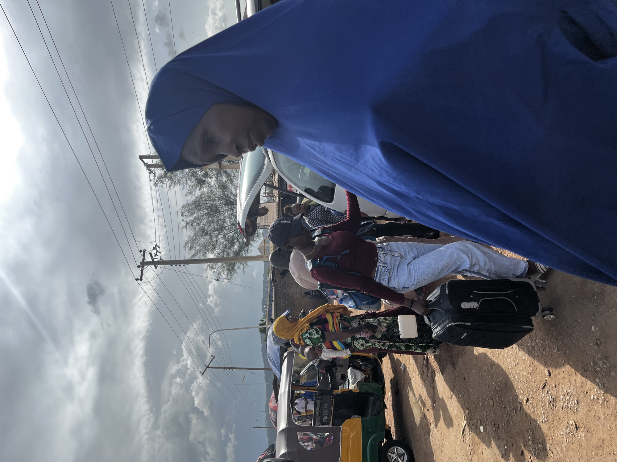 The image depicts a street scene in Rafin Par, Nigeria, on a cloudy day. Several people are visible, some appearing to be waiting or preparing for travel.
In the foreground, a person wearing a large blue hijab is partially visible, their face turned downwards and to the side. Behind them, a line of people are gathered near vehicles. One person, wearing a maroon long-sleeved shirt and light blue jeans, is handling a black suitcase with wheels. Another individual in a vibrant yellow and multi-colored patterned headscarf and dress is standing near a three-wheeled auto-rickshaw, commonly known as a "Keke Napep." The auto-rickshaw is a mix of brown and green, with visible text that includes "PM 1103A" on the side.
Other individuals are seen in the background, some appearing to be seated in or near vehicles. The presence of luggage, the auto-rickshaw, and the gathering of people suggest a transportation hub or a moment of departure or arrival. The sky is overcast with dramatic clouds, indicating a potentially rainy or unsettled weather condition. The ground is a mixture of dirt and gravel. Power lines and utility poles are visible in the background, suggesting an urban or semi-urban setting. The overall mood seems to be one of transit and activity.