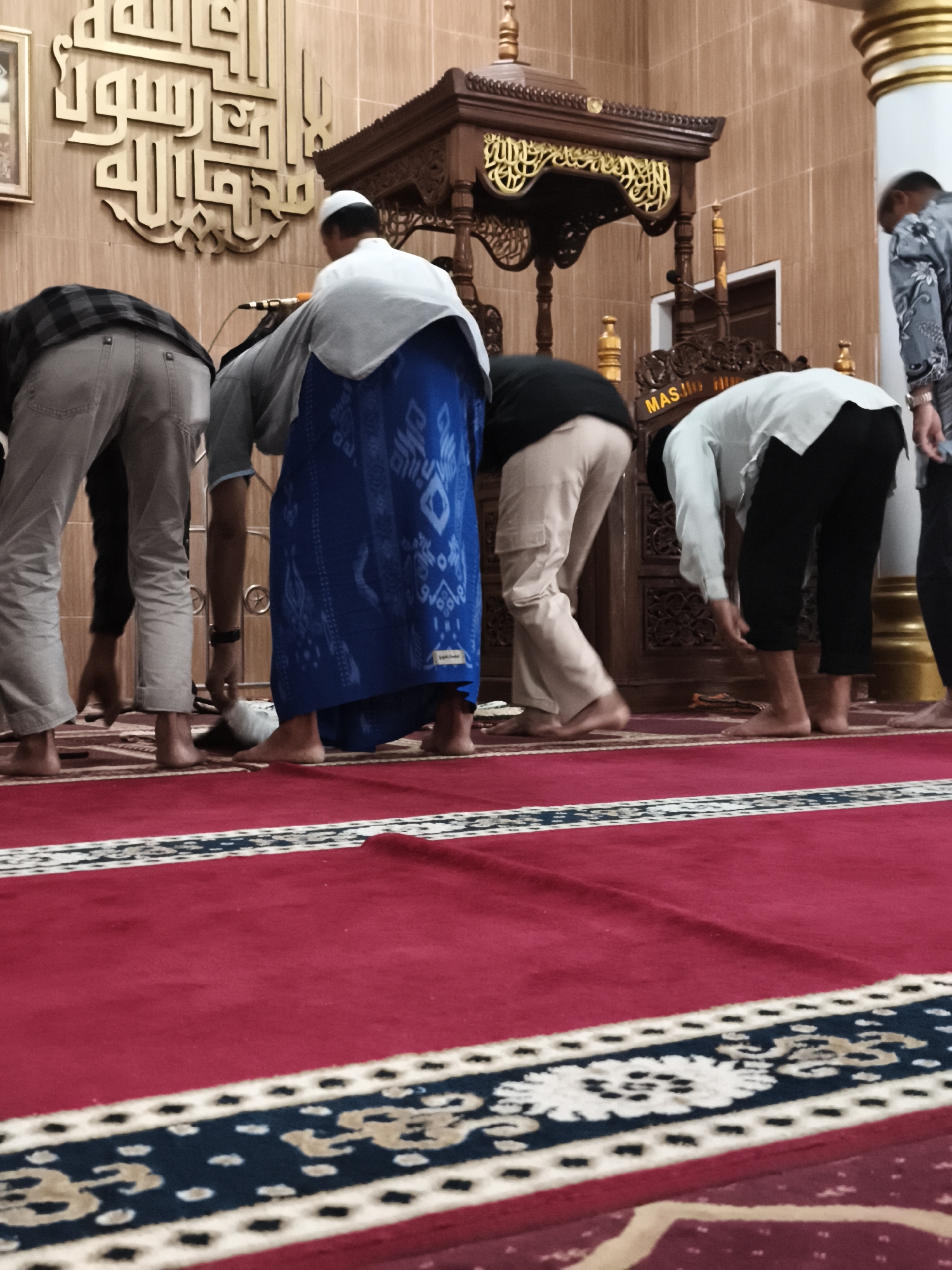 This is a medium-shot, eye-level photograph taken indoors, likely in a mosque, with the location specified as Tonasa, Indonesia. The scene depicts at least four men engaged in prayer, indicated by their bowed postures. The floor is covered with a red carpet featuring a decorative border with intricate patterns in blue and cream.
In the foreground, the legs and lower torsos of three men are visible. The man on the far left wears light-colored trousers and a checkered shirt, with his back to the camera. Next to him, a man wears light-colored pants and a grey shirt, with his head bowed. The third man from the left is wearing a blue sarong, with his back to the camera and head bowed.
In the mid-ground, a wooden pulpit with ornate carvings stands prominently. Behind the pulpit, a wall with a patterned texture is visible, adorned with large, golden Arabic calligraphy. To the right of the pulpit, another man in a white shirt and black pants is in a prayer posture, with his head bowed towards the ground. Further to the right, a man wearing a patterned shirt is partially visible. The lighting appears to be warm and ambient.