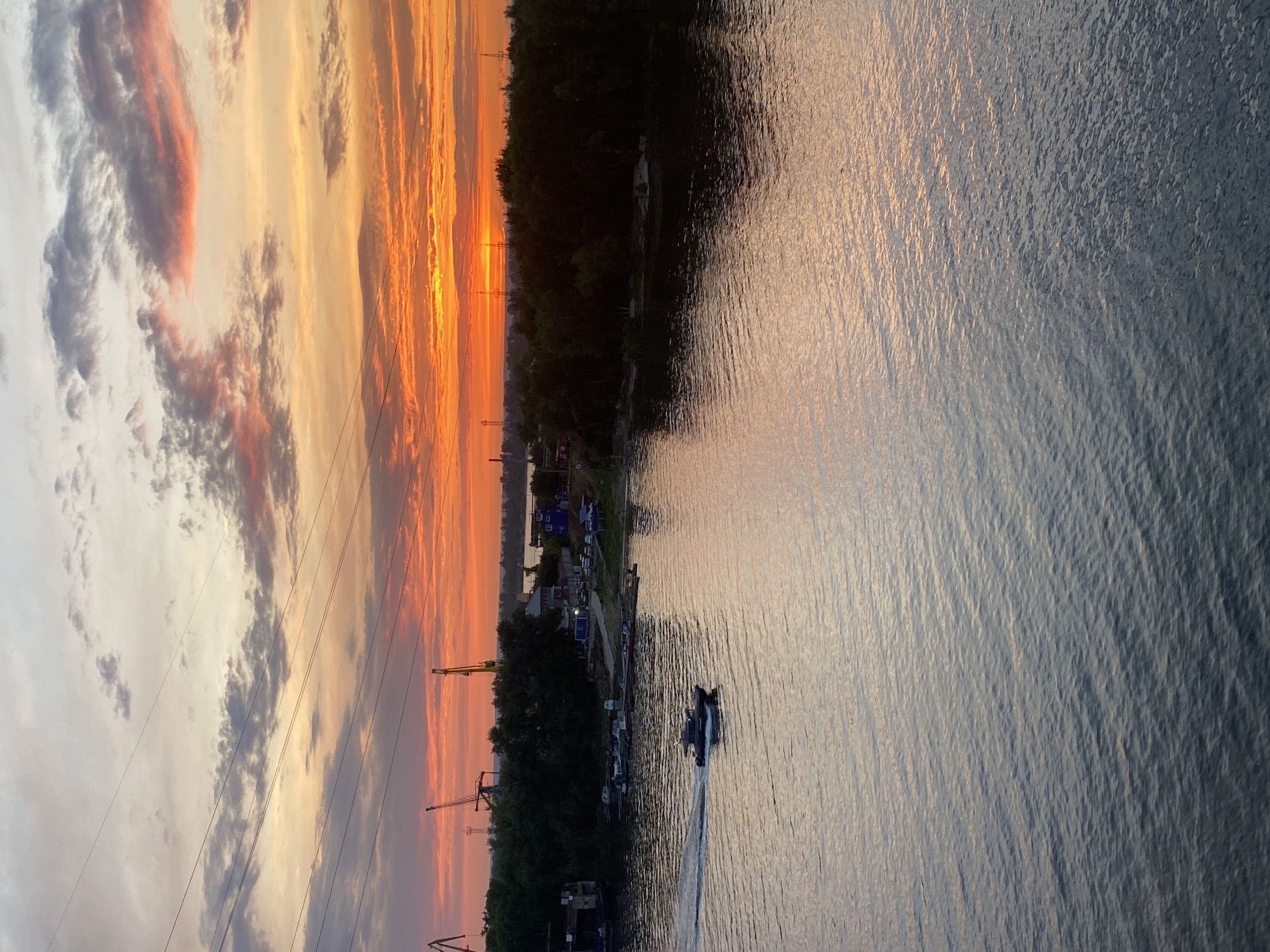 This image depicts a river scene at sunset in Samara, Russia. The sky is ablaze with vibrant orange and pink hues, characteristic of a sunset, with scattered clouds. Overhead, power lines stretch across the frame. In the distance, along the riverbank, industrial cranes are visible, suggesting a port or industrial area. A lush green treeline forms a dark silhouette against the colorful sky. On the water, a small boat is moving, leaving a wake. The river itself is calm, reflecting the colors of the sky. The overall impression is one of natural beauty and industrial activity coexisting.