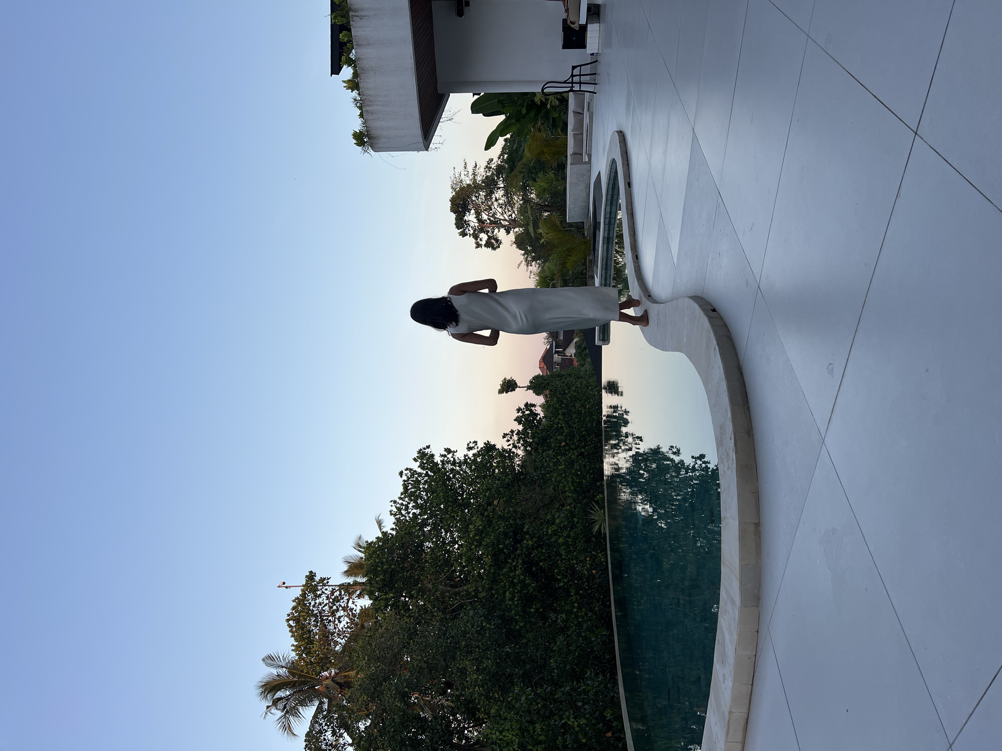 A woman in a light-colored dress stands with her back to the camera, facing away from a curved infinity pool and a modern white building. The pool's surface reflects the trees and the sky. Lush green trees and palm fronds surround the property. The sky is a clear blue with a hint of pink near the horizon, suggesting either dawn or dusk. The scene appears to be a resort or private villa in the tropical environment of Ubud District, Indonesia, characterized by its abundant foliage and serene atmosphere.