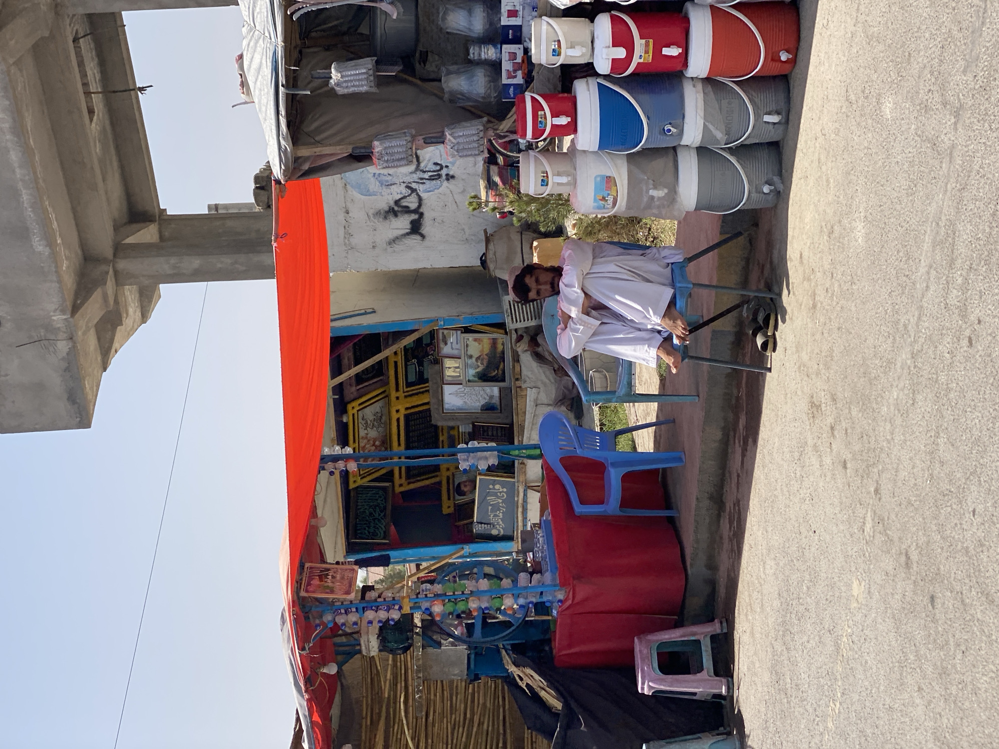 A man in traditional Afghan clothing, a white shalwar kameez and a cap, sits on a blue plastic chair at a roadside stall in Kandahar, Afghanistan. He is looking forward with a neutral expression, his arms crossed. The stall is decorated with a bright red awning and displays various items, including framed pictures, bottles of water, and large water coolers. To the left of the man, a pink plastic stool is visible. The ground is paved with asphalt, and a building with concrete pillars stands in the background. The scene appears to be an outdoor market or vendor setup.